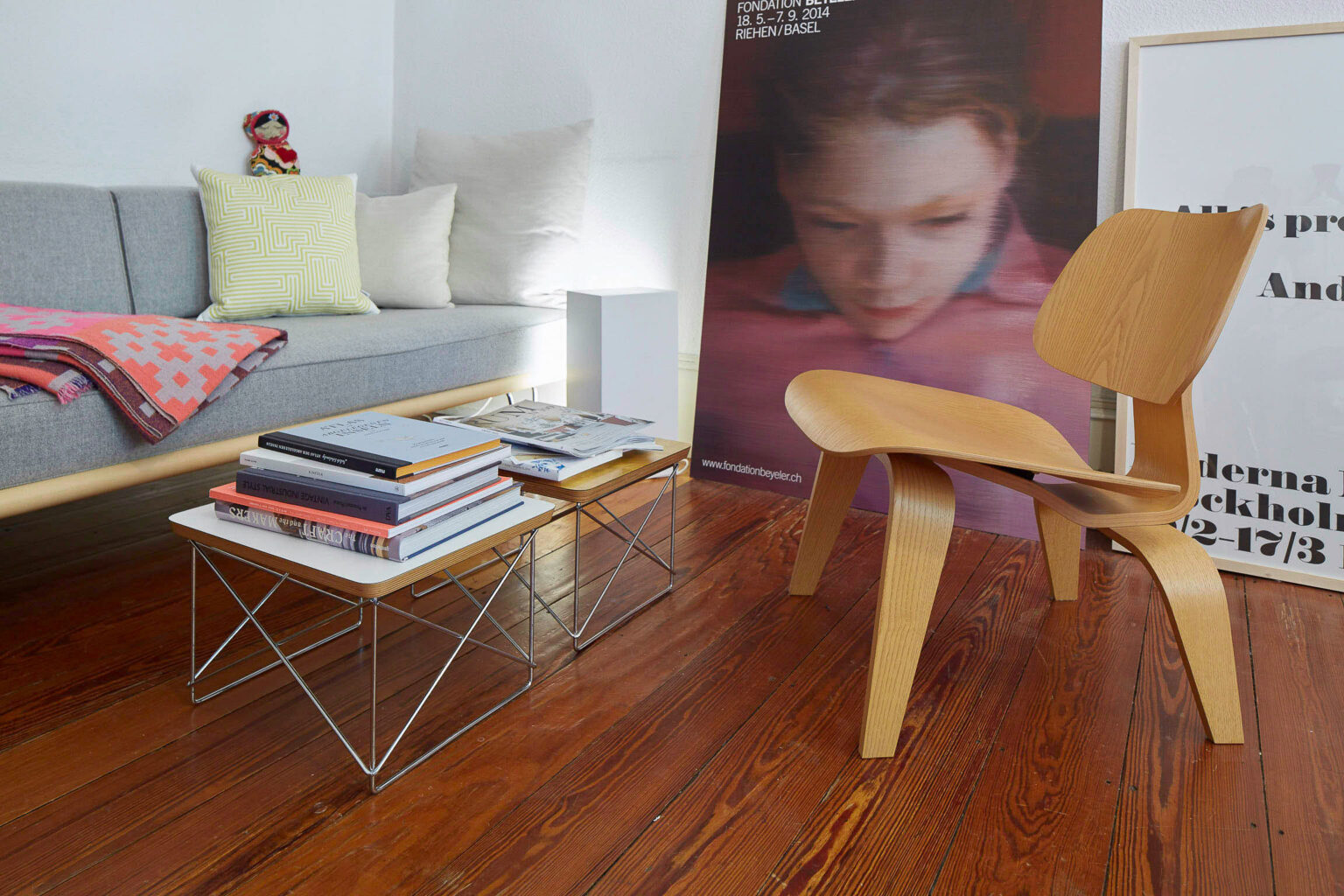 Ein Wohnzimmer mit einem grauen Sofa, auf dem mehrere Kissen und eine bunte Decke liegen. Vor dem Sofa stehen zwei niedrige Tische aus Holz und Metall, die mit einem Stapel Bücher und Zeitschriften bedeckt sind. Rechts vom Sofa steht ein moderner Holzstuhl. An der Wand hinter dem Stuhl lehnen zwei große Kunstplakate, eines zeigt das Bild eines jungen Mädchens, das andere Text. Der Raum hat einen Holzboden.