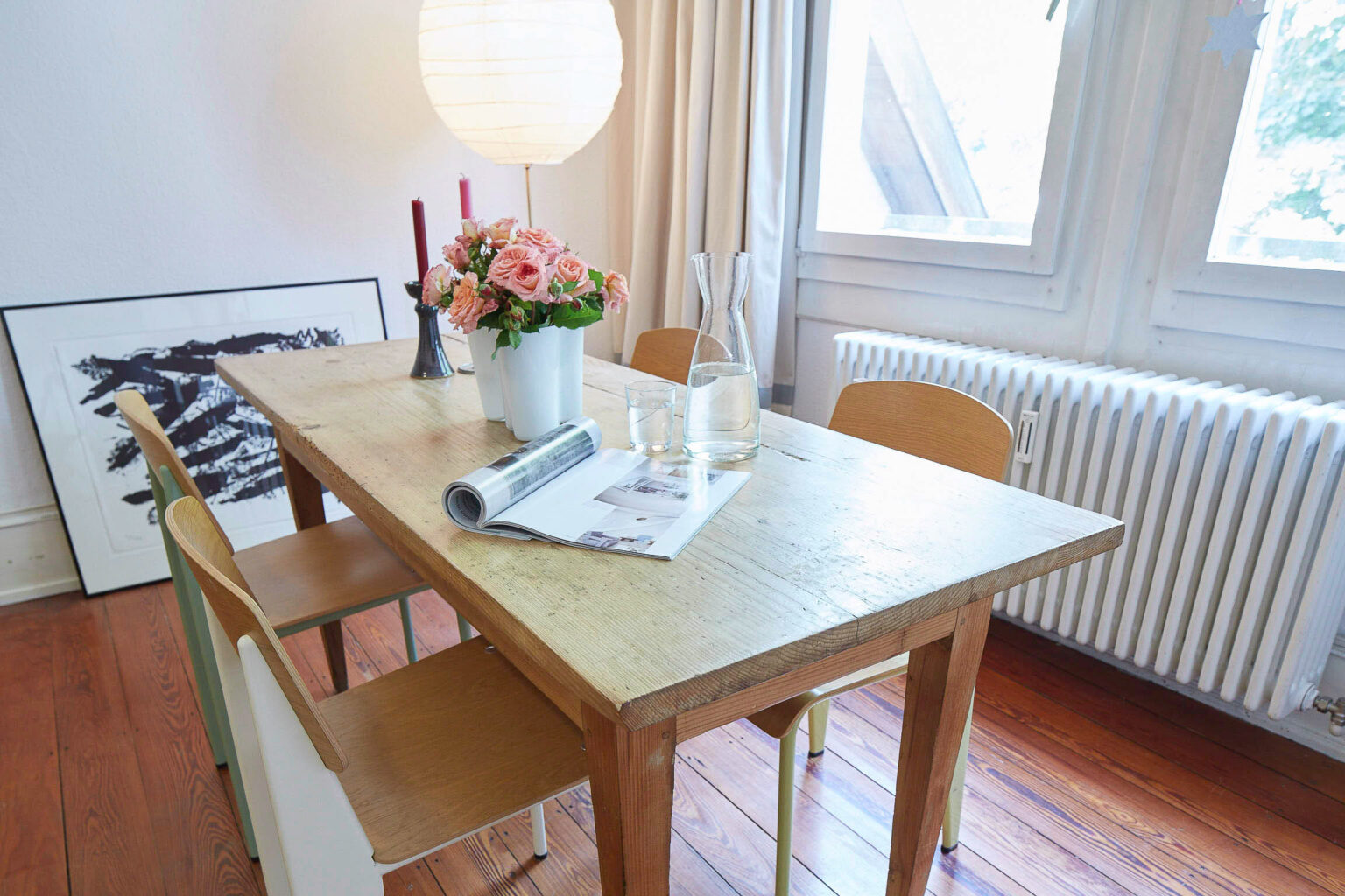 Ein Essbereich mit einem rechteckigen Holztisch und vier Holzstühlen. Auf dem Tisch stehen eine Vase mit rosa Rosen, zwei Kerzenhalter mit roten Kerzen, eine Glaskaraffe mit Wasser und ein Glas. Ein aufgeschlagenes Magazin liegt auf dem Tisch. Im Hintergrund hängt eine große Papierlaterne von der Decke, und durch die Fenster rechts fällt Tageslicht in den Raum. Auf dem Boden lehnt ein gerahmtes Kunstwerk an der Wand. Der Raum hat einen Holzboden und weiße Heizkörper unter den Fenstern.