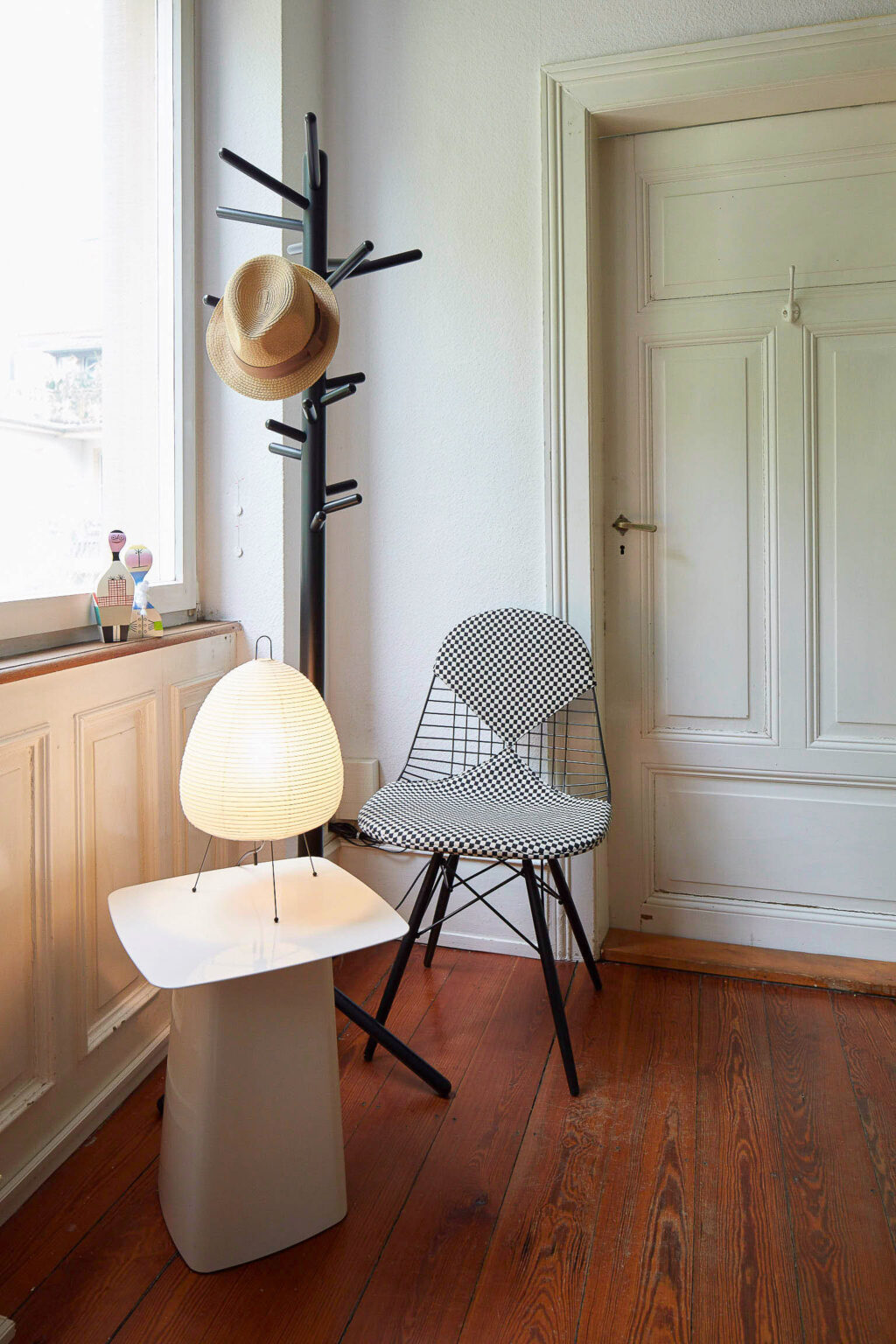 Ein heller Eingangsbereich mit einem schwarz-weiß karierten Stuhl und einem schwarzen Garderobenständer, an dem ein Strohhut hängt. Vor dem Stuhl steht ein kleiner, moderner Tisch mit einer weißen Oberfläche, auf dem eine beleuchtete Tischlampe in Form einer traditionellen japanischen Laterne steht. Neben dem Tisch, auf der Fensterbank, stehen zwei kleine dekorative Figuren. Der Raum hat einen Holzboden und eine weiße Tür.