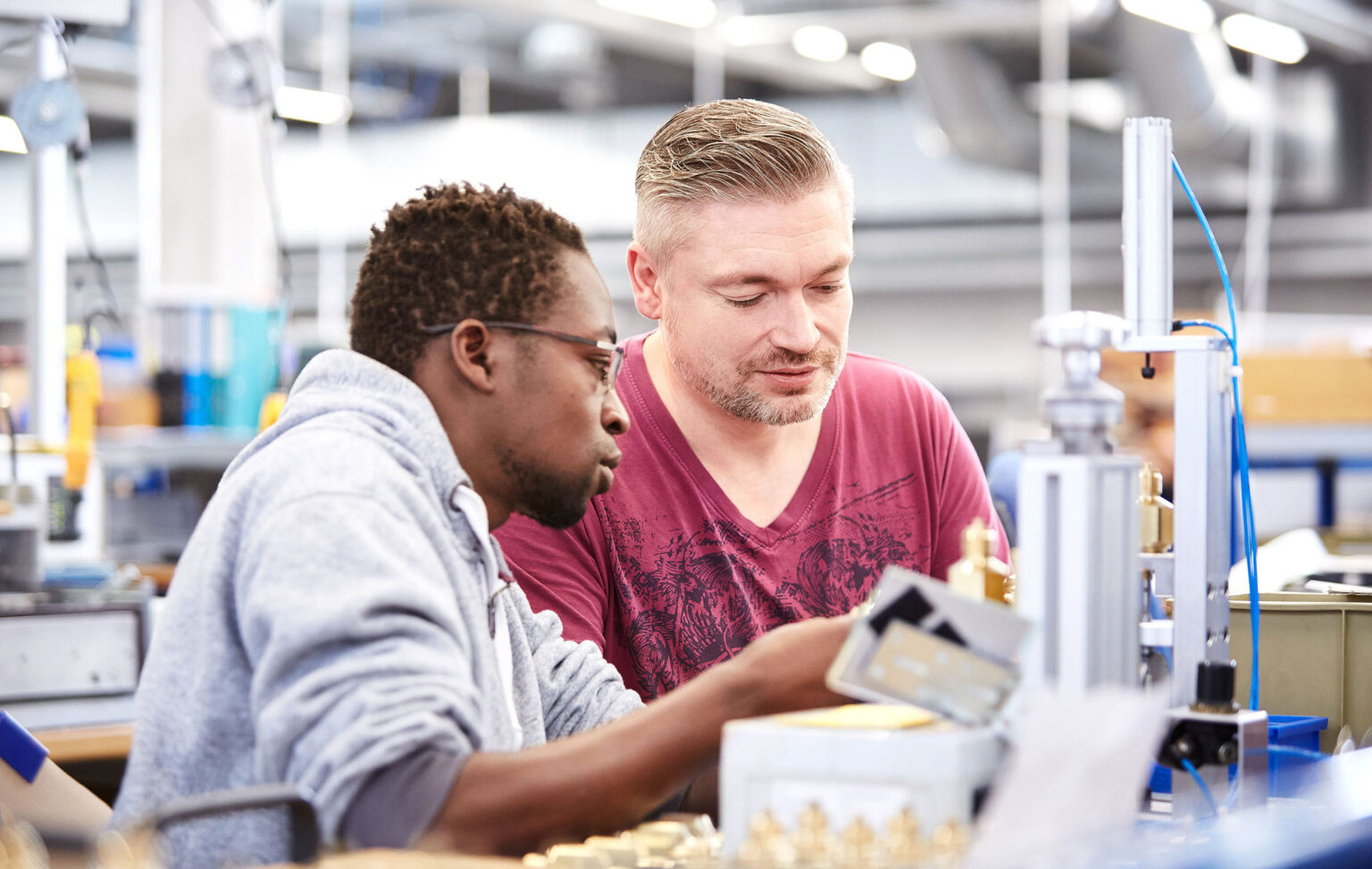 Zwei Männer arbeiten konzentriert an einer Maschine in einer industriellen Umgebung. Der Mann links trägt eine Brille und einen grauen Kapuzenpullover, der Mann rechts hat kurzes blondes Haar und trägt ein rotes T-Shirt. Beide betrachten ein Bauteil in ihren Händen.