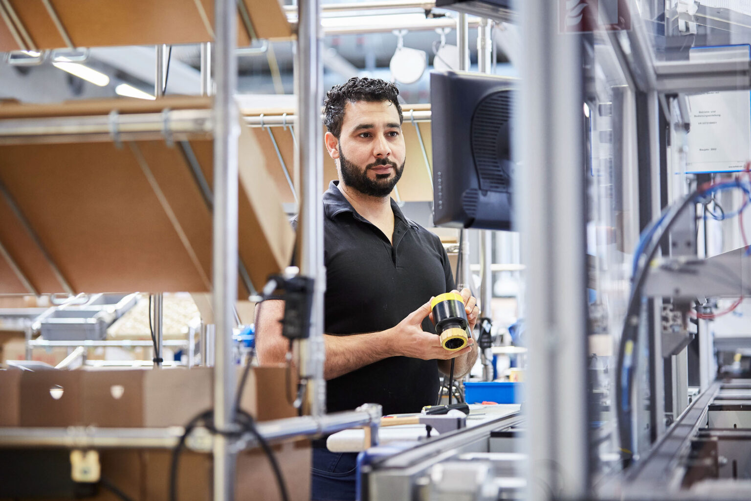 Ein Mann mit schwarzem Bart und lockigem Haar trägt ein schwarzes Poloshirt. Er steht in einer industriellen Umgebung und hält ein gelbes Gerät in den Händen, während er auf einen Monitor vor sich schaut. Um ihn herum sind Metallregale und verschiedene technische Geräte zu sehen.
