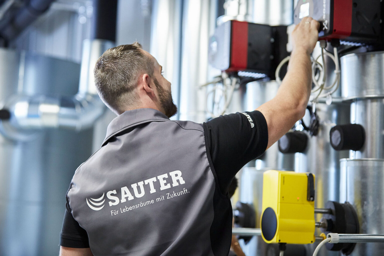 Ein Mann mit kurzem Haar und Bart trägt eine graue Weste mit der Aufschrift 'SAUTER Für Lebensräume mit Zukunft' und ein schwarzes Poloshirt. Er arbeitet an einer technischen Anlage in einem Industriegebäude, umgeben von Rohren und Geräten. Der Mann ist von hinten zu sehen, wie er eine Hand zu einem der Geräte ausstreckt.