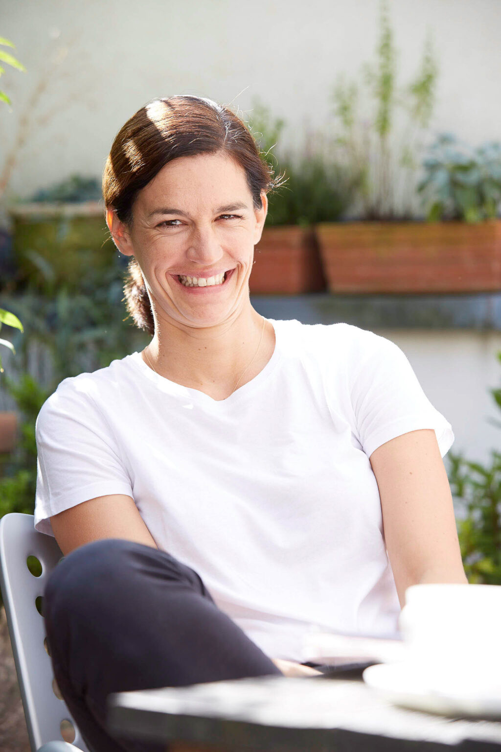Eine Frau mit braunen, nach hinten gebundenen Haaren sitzt lächelnd draußen an einem Tisch. Sie trägt ein weißes T-Shirt und schwarze Hosen. Im Hintergrund sind Pflanzen und Blumenkästen zu sehen.