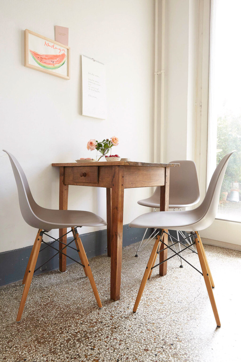 Ein kleiner, gemütlicher Essbereich mit einem rustikalen Holztisch und zwei modernen Stühlen mit grauen Sitzflächen und Holzbeinen. Auf dem Tisch stehen eine Vase mit rosafarbenen Blumen und eine Schale mit roten Früchten. An der weißen Wand hinter dem Tisch hängen ein Kalender und ein gerahmtes Bild einer Wassermelone. Der Boden ist mit kleinen, unregelmäßigen Fliesen bedeckt und rechts ist eine große Fensterfront zu sehen, die viel Tageslicht hereinlässt und einen Blick auf die grüne Außenwelt bietet."