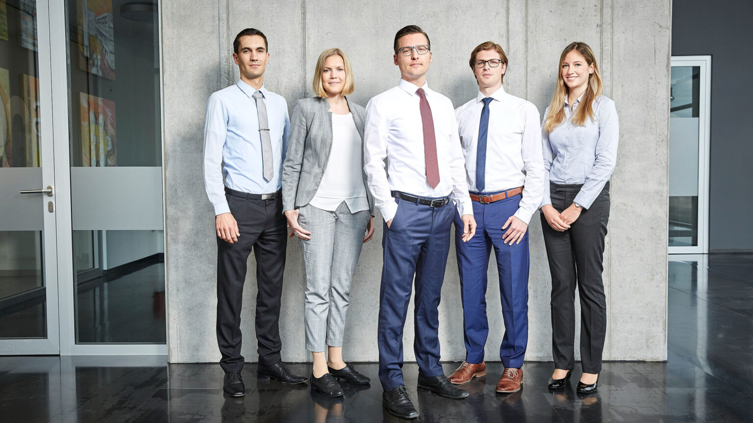 Fünf Personen in Businesskleidung stehen nebeneinander vor einer Betonwand. Von links nach rechts tragen sie folgende Kleidung: ein Mann in einem hellblauen Hemd und einer schwarzen Hose mit grauer Krawatte, eine Frau in einem grauen Anzug mit weißer Bluse, ein Mann in einem weißen Hemd und einer dunkelblauen Hose mit roter Krawatte, ein Mann in einem weißen Hemd und einer blauen Hose mit blauer Krawatte und ein brauner Gürtel, und eine Frau in einer hellblauen Bluse und einer schwarzen Hose. Im Hintergrund ist ein modernes Bürogebäude mit Glastüren und Gemälden an den Wänden zu sehen.