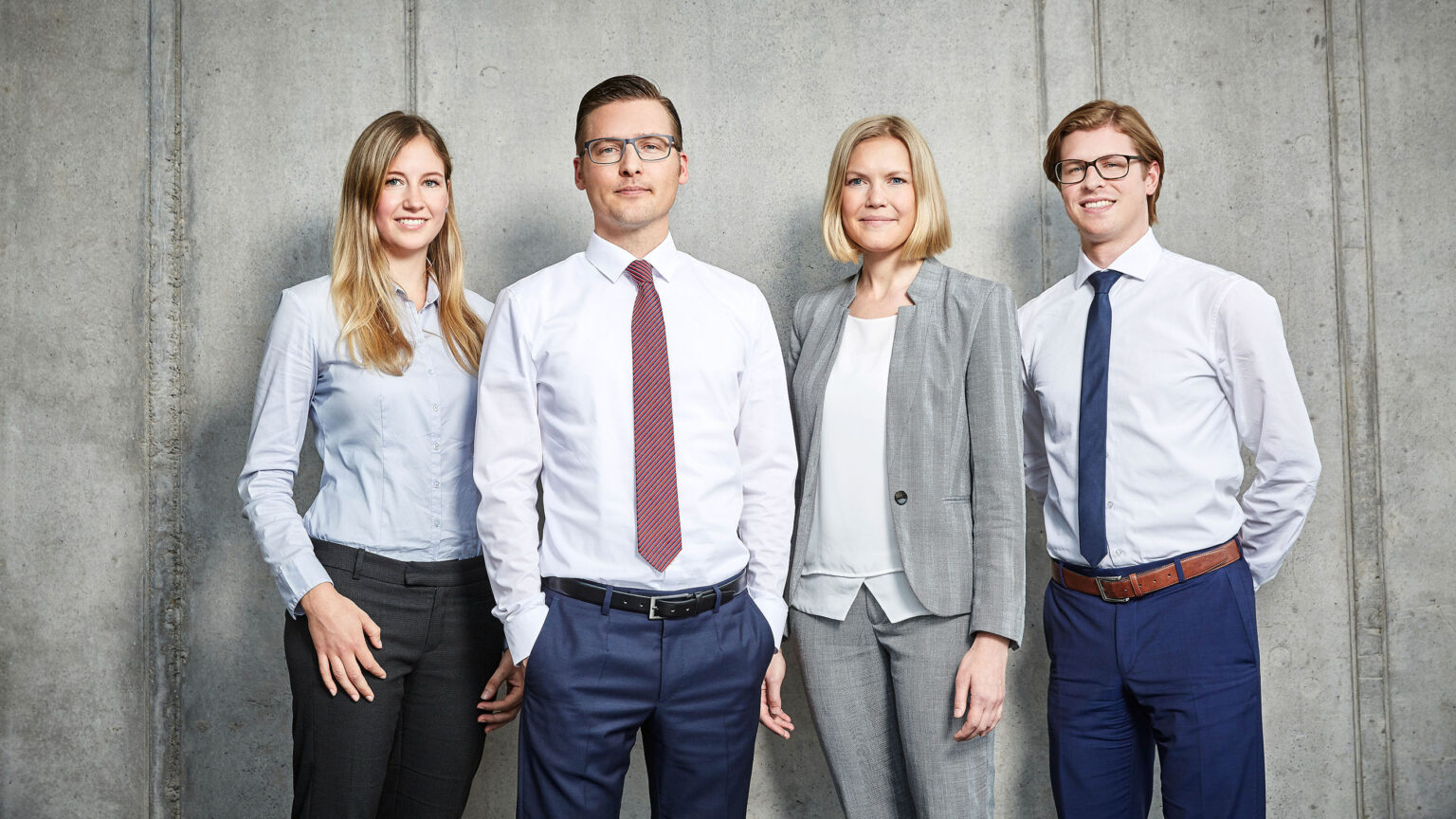 Vier Personen in Businesskleidung stehen vor einer Betonwand. Von links nach rechts tragen sie folgende Kleidung: eine Frau in einer hellblauen Bluse und dunkler Hose, ein Mann in einem weißen Hemd und dunkelblauer Hose mit roter Krawatte, eine Frau in einem grauen Anzug mit weißer Bluse, und ein Mann in einem weißen Hemd und blauer Hose mit dunkelblauer Krawatte und braunem Gürtel. Alle vier Personen schauen freundlich in die Kamera.