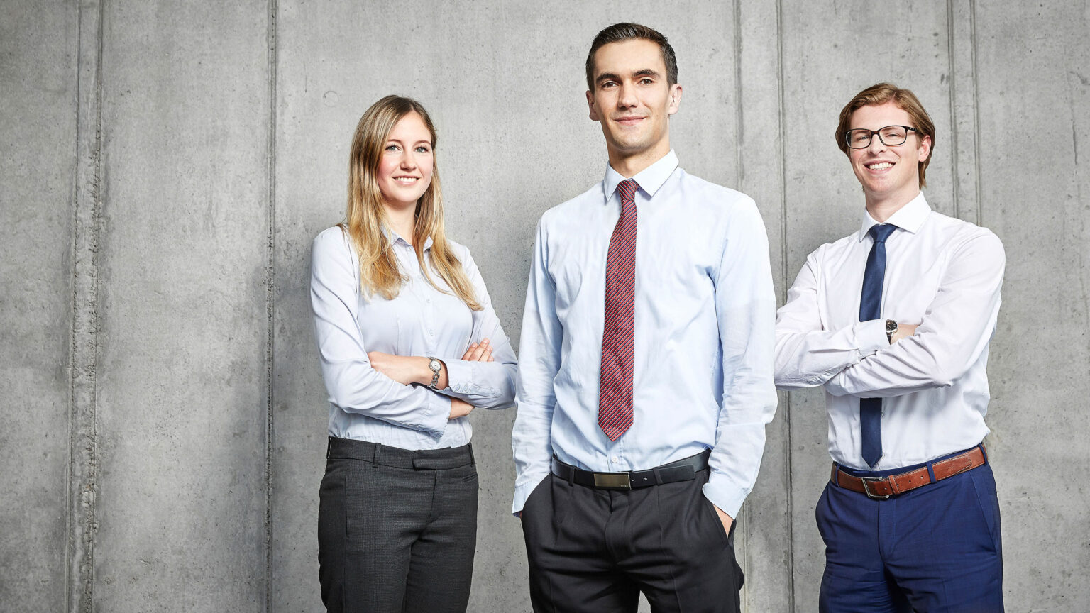 Drei Personen in Businesskleidung stehen vor einer Betonwand. Von links nach rechts tragen sie folgende Kleidung: eine Frau in einer hellblauen Bluse und einer dunklen Hose mit verschränkten Armen, ein Mann in einem hellblauen Hemd und einer schwarzen Hose mit roter Krawatte und Händen in den Hosentaschen, und ein Mann in einem weißen Hemd und einer blauen Hose mit dunkelblauer Krawatte und verschränkten Armen. Alle drei Personen lächeln in die Kamera.