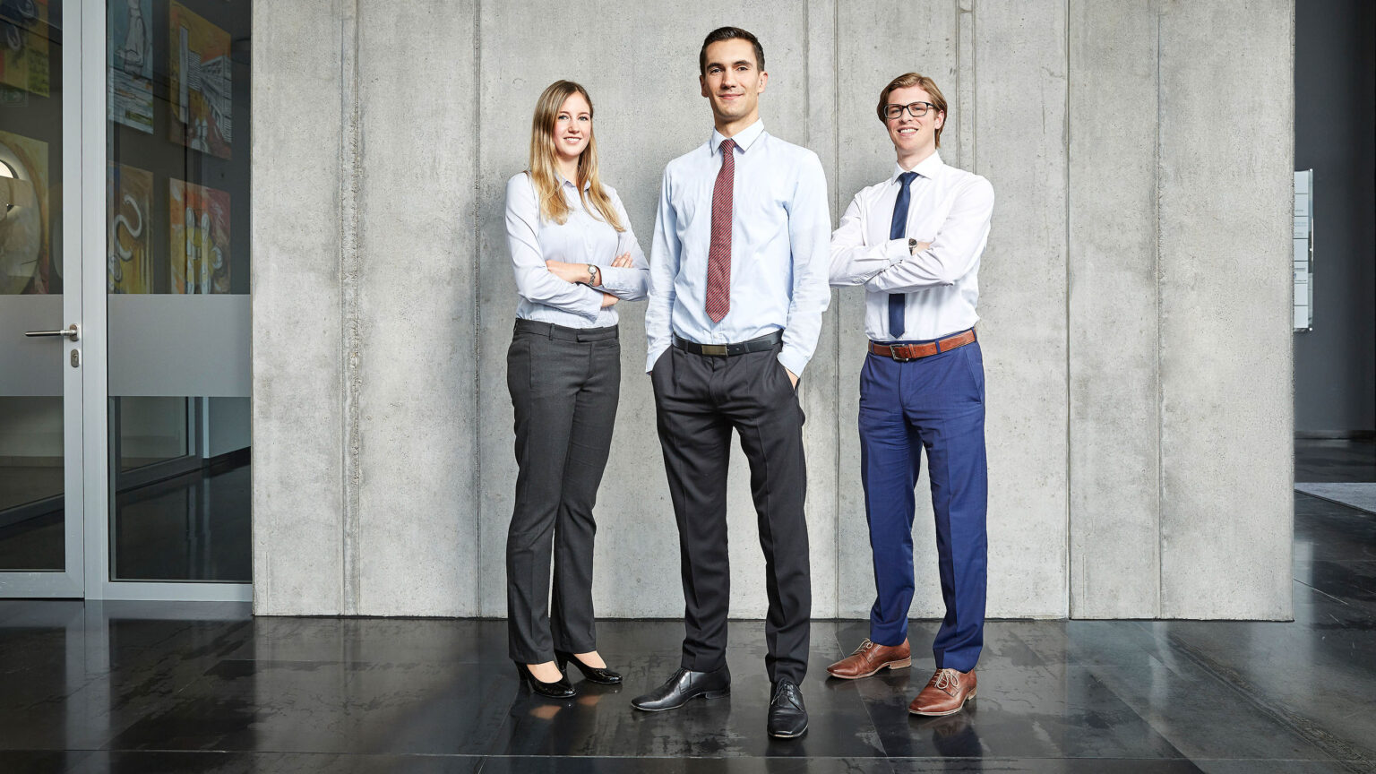 Drei Personen in Businesskleidung stehen vor einer Betonwand. Von links nach rechts tragen sie folgende Kleidung: eine Frau in einer hellblauen Bluse und einer dunklen Hose mit verschränkten Armen, ein Mann in einem hellblauen Hemd und einer schwarzen Hose mit roter Krawatte und Händen in den Hosentaschen, und ein Mann in einem weißen Hemd und einer blauen Hose mit dunkelblauer Krawatte und verschränkten Armen. Im Hintergrund ist eine Glastür zu sehen, die in einen modernen Büroraum mit Gemälden an den Wänden führt.