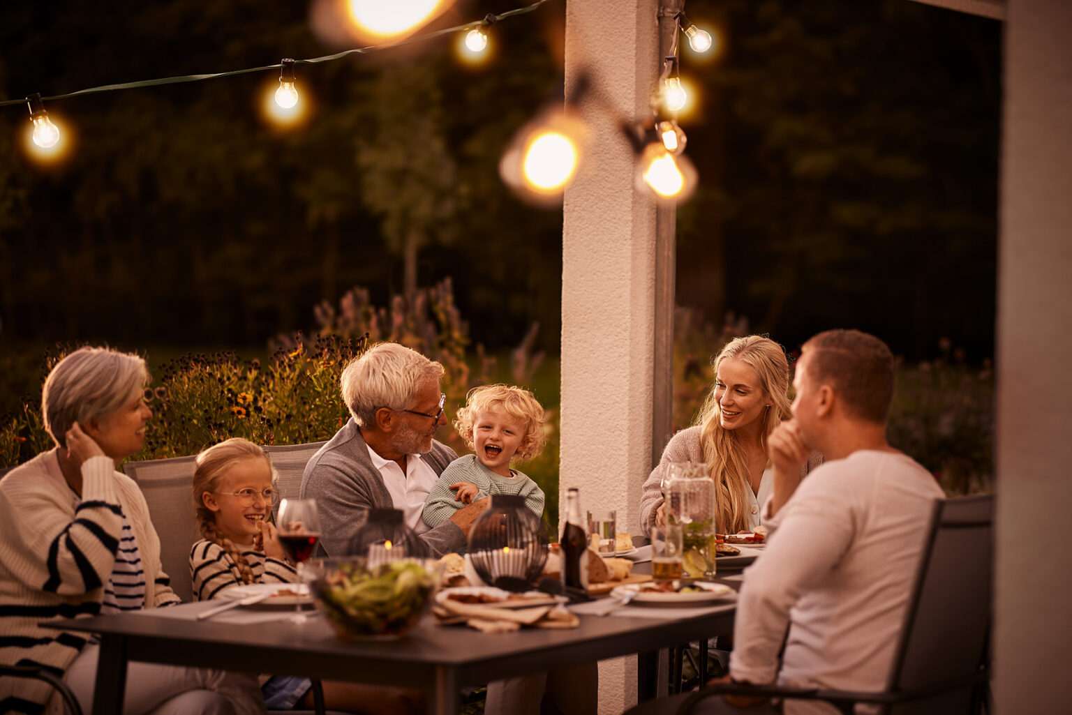 Eine mehrere Generationen umfassende Familie sitzt am Abend um einen Tisch im Freien, umgeben von der sanften Beleuchtung von Lichterketten. Ein älterer Herr lacht herzlich mit einem kleinen blonden Kind, das auf dem Schoß einer jungen Frau sitzt. Eine andere Frau, möglicherweise die Großmutter, und ein Mädchen mit Brille lächeln ebenfalls, während ein junger Mann dem Gespräch zuhört. Der Tisch ist mit Gläsern und Tellern für ein gemeinsames Essen gedeckt, und die warme Beleuchtung trägt zu einer gemütlichen und intimen Atmosphäre bei. Im Hintergrund ist ein bisschen von einem gepflegten Garten und dem dunklen Abendhimmel zu erkennen.