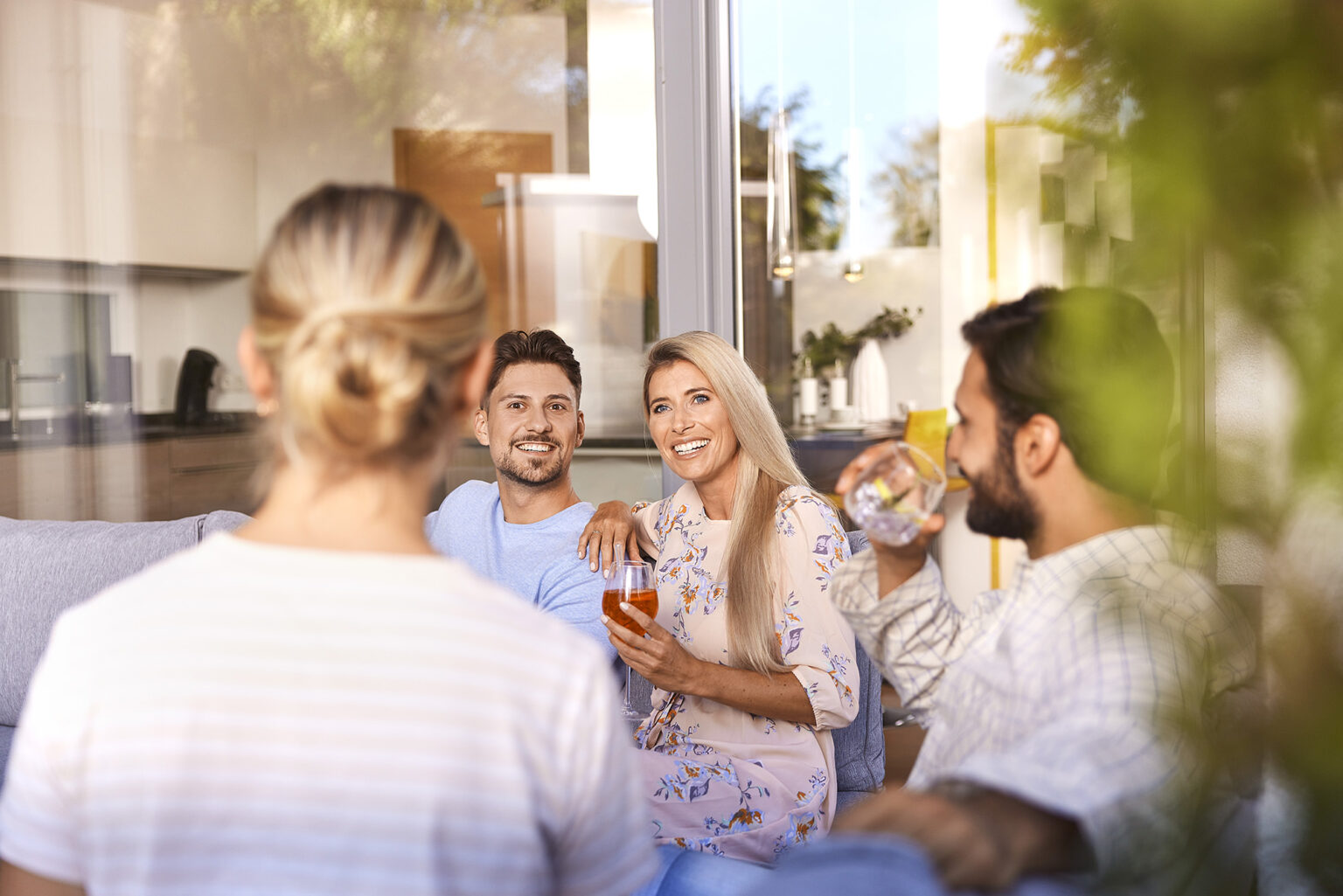 Ein fröhlicher Freundeskreis genießt gemeinsam die Zeit auf einer sonnigen Terrasse. Die Aufnahme zeigt vier Personen von hinten und von der Seite, wobei eine blonde Frau in einem geblümten Kleid, die in die Kamera lächelt, im Vordergrund steht. Sie hält ein Glas mit einem rosafarbenen Getränk. Ein Mann mit dunklem Haar und blauem T-Shirt sitzt neben ihr und lächelt, während er in die Kamera blickt. Eine weitere Person ist im Profil zu sehen, trinkt aus einem Weinglas und der Blick geht auf eine Person, die von hinten zu sehen ist. Im Hintergrund erkennt man die stilvolle Inneneinrichtung durch die Glasscheiben des modernen Hauses.