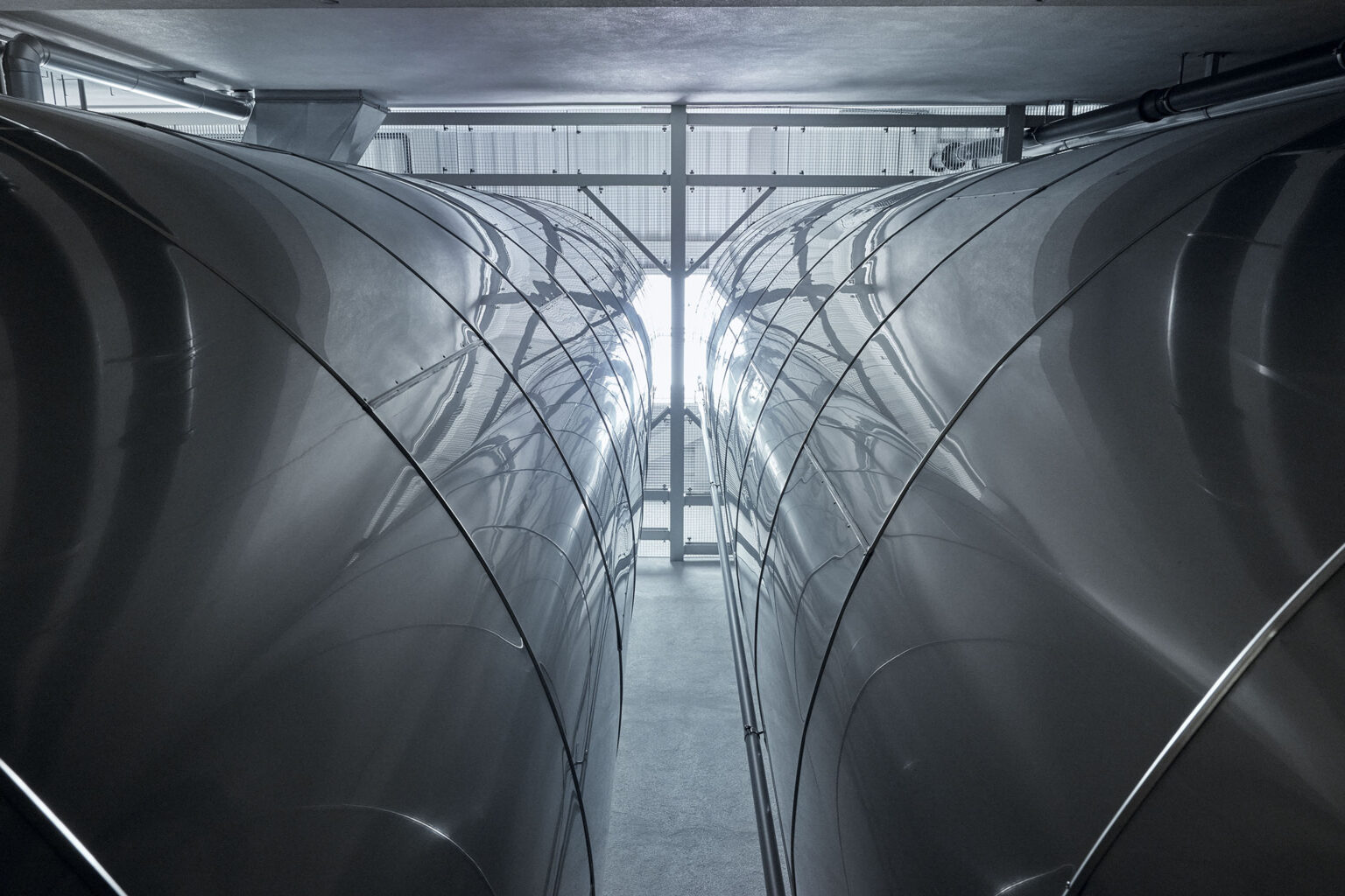 Ein Blick zwischen zwei große, glänzende Edelstahlbehälter in einer industriellen Anlage. Die Behälter sind symmetrisch angeordnet und reflektieren das Licht, was ihre glatte Oberfläche betont. Am Ende des Ganges zwischen den Behältern ist ein Fenster zu sehen, das Tageslicht hereinlässt und den Raum beleuchtet. Die gesamte Umgebung wirkt sauber und modern.
