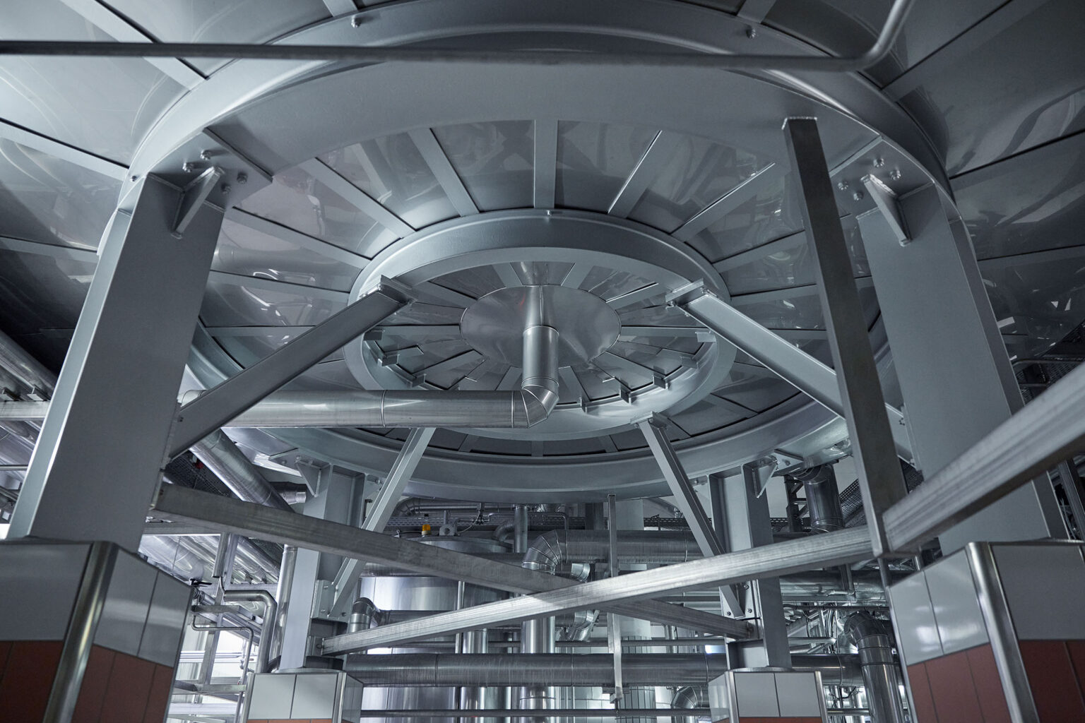 Innenansicht einer industriellen Anlage, die eine große, runde Stahlkonstruktion zeigt. Die Konstruktion wird von mehreren massiven Stahlträgern gestützt und ist Teil eines komplexen Rohr- und Leitungssystems. Die glänzende Oberfläche der Struktur reflektiert das Licht, was die moderne und sterile Umgebung der Anlage betont.