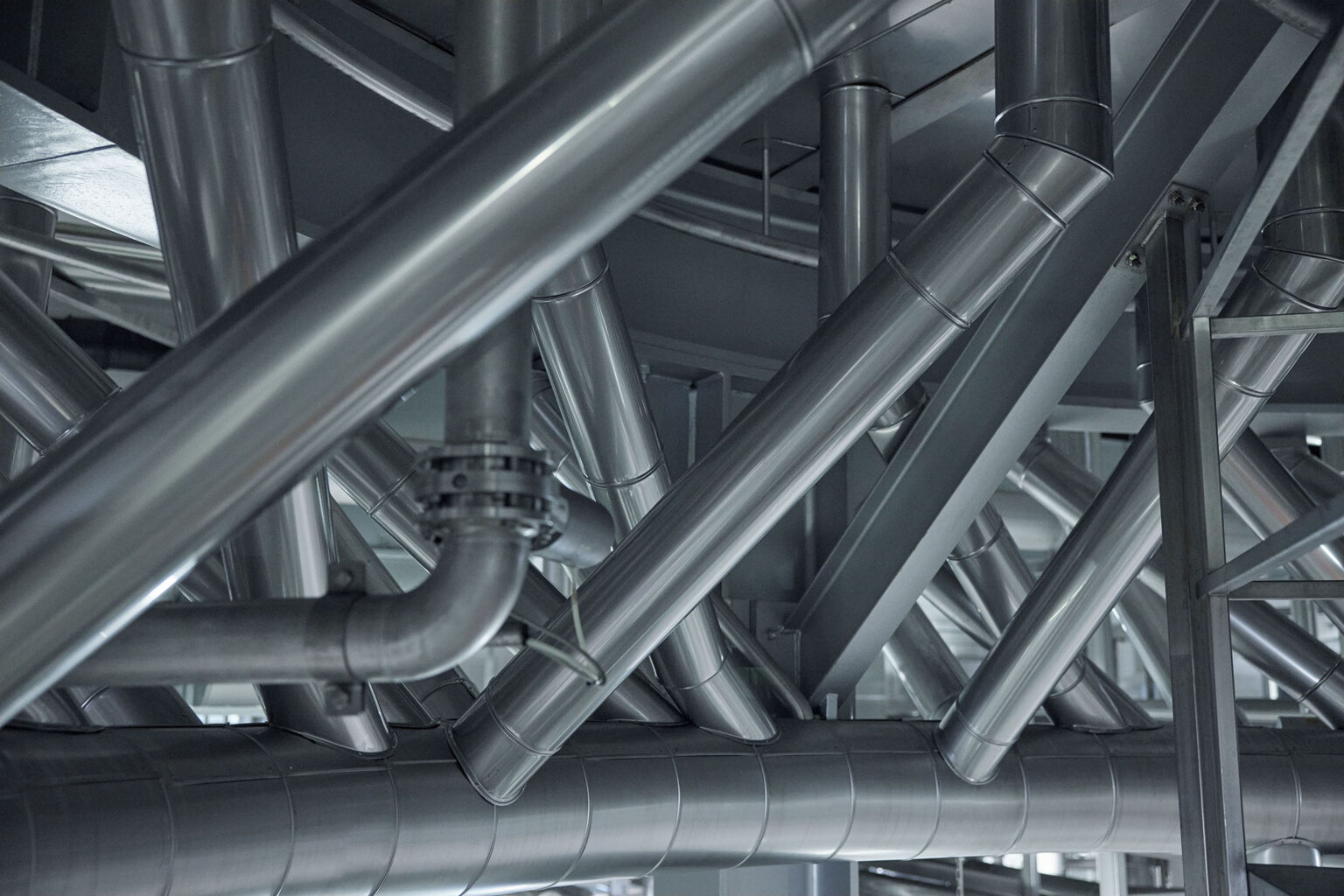 Ein komplexes Netzwerk aus glänzenden Edelstahlrohren und -leitungen in einer industriellen Anlage. Die Rohre verlaufen in verschiedenen Winkeln und Richtungen und sind stabil an der Decke und an den Wänden montiert. Die gesamte Struktur wirkt robust und gut organisiert, was auf eine moderne und effiziente industrielle Umgebung hinweist. Das glänzende Metall reflektiert das Licht und betont die saubere und gut gewartete Anlage.