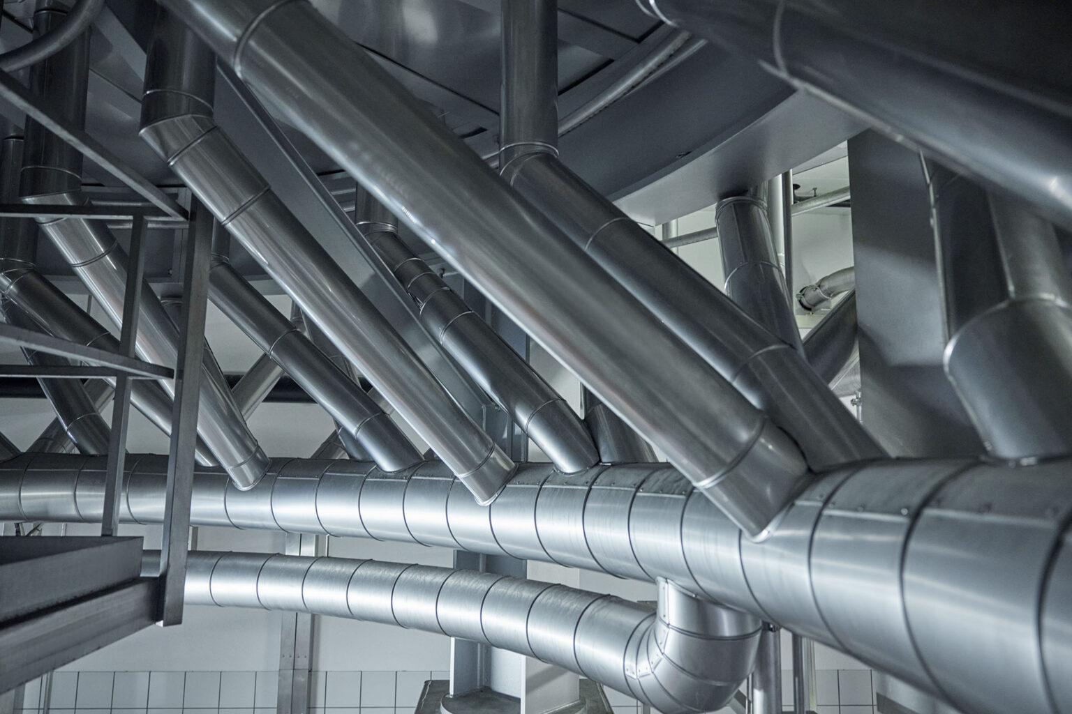 Ein komplexes Netzwerk von glänzenden Edelstahlrohren und Leitungen in einer industriellen Anlage. Die Rohre sind in verschiedenen Winkeln und Richtungen angeordnet und durch eine stabile Metallstruktur gestützt. Die gesamte Umgebung wirkt sauber und modern, und die Rohre reflektieren das Licht, was die hochwertige Verarbeitung und das effiziente Design der Anlage betont.