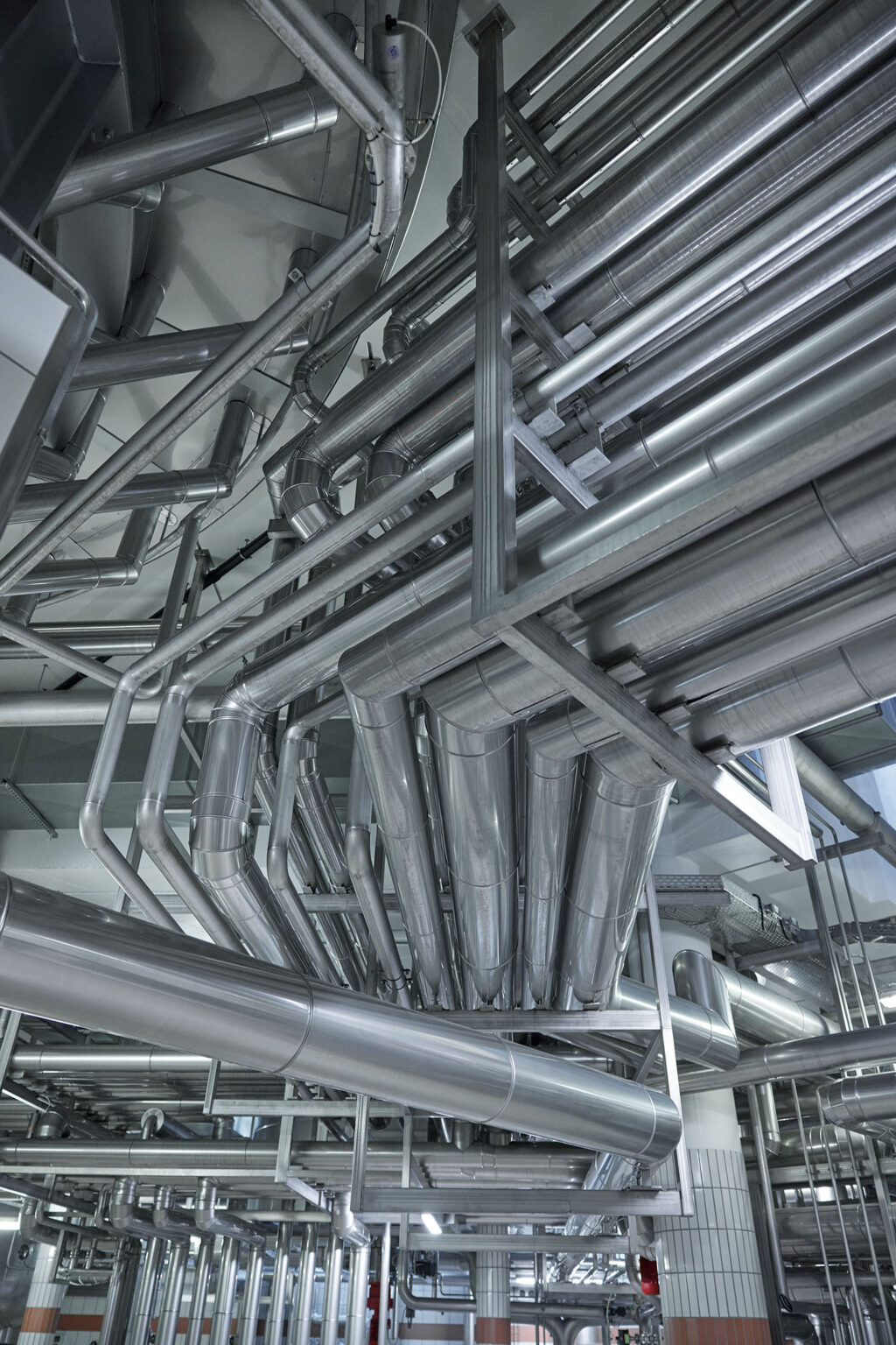 Netzwerk aus Edelstahlrohren und -leitungen in einer industriellen Anlage, die sich komplex und verschlungen in verschiedenen Richtungen erstrecken. Die Rohre und Leitungen verlaufen über Kopf und sind durch Rahmenstrukturen stabilisiert. Die glänzende Oberfläche der Rohre reflektiert das Licht, was die moderne und saubere Umgebung der Anlage unterstreicht