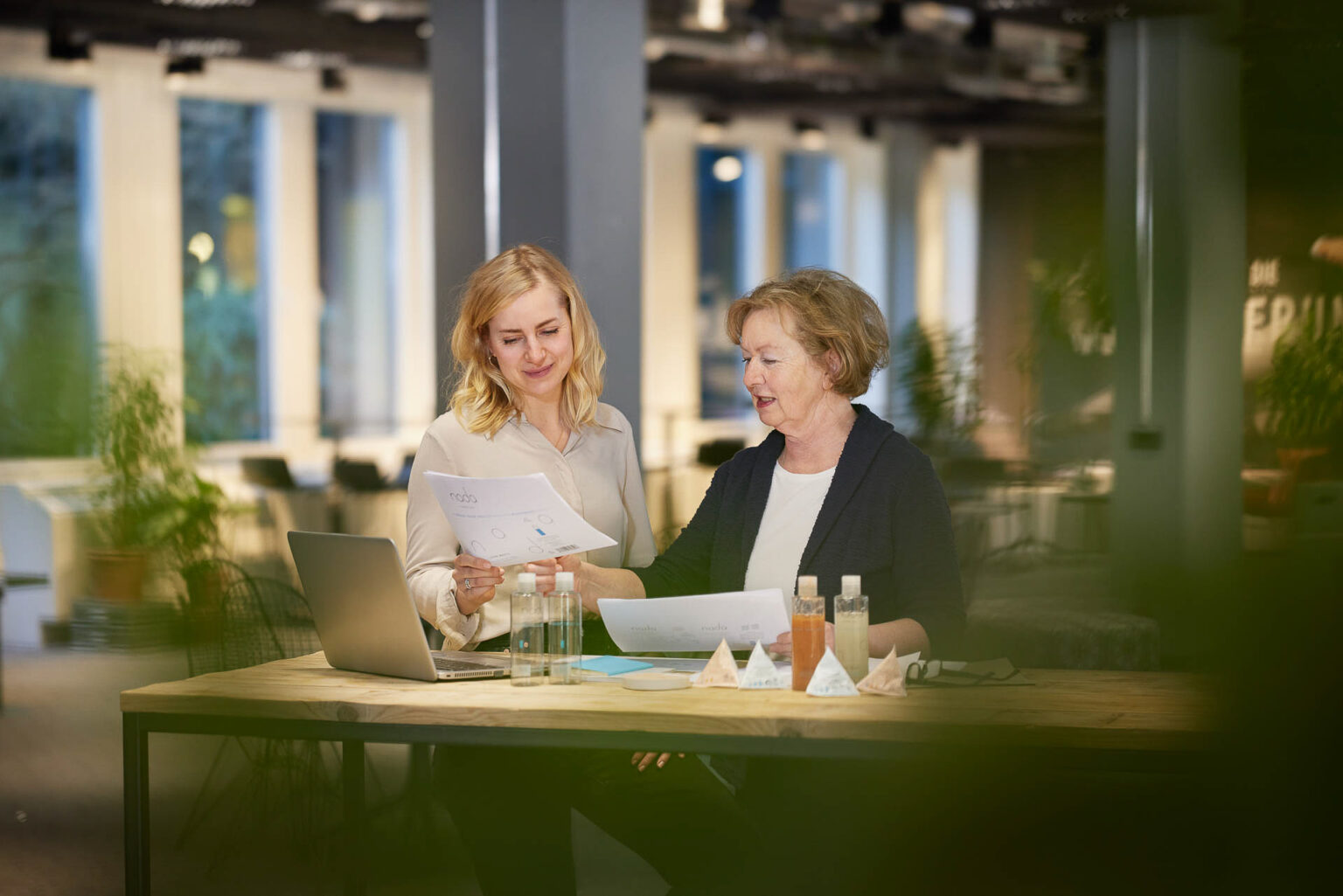 Zwei Frauen sitzen an einem Tisch in einem modernen, hellen Raum. Die Frau links hat schulterlanges blondes Haar und trägt eine helle Bluse. Sie hält ein Blatt Papier in der Hand und lächelt. Die Frau rechts hat kurzes blondes Haar und trägt eine dunkelblaue Jacke. Sie zeigt auf das Papier in ihrer Hand. Auf dem Tisch stehen ein Laptop, mehrere Flaschen und einige dreieckige Verpackungen.
