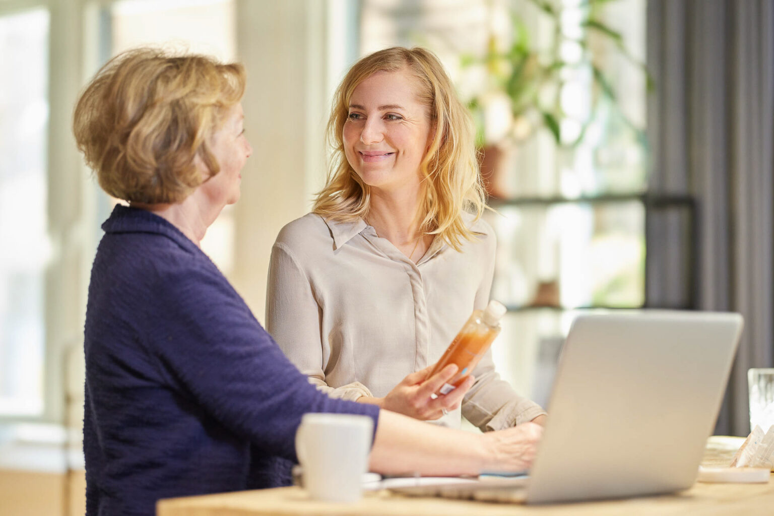 Zwei Frauen unterhalten sich in einem hellen Raum. Die Frau auf der linken Seite hat kurzes blondes Haar und trägt eine dunkelblaue Jacke. Die Frau auf der rechten Seite hat schulterlanges blondes Haar und trägt eine helle Bluse. Sie hält eine Flasche mit einer orangefarbenen Flüssigkeit in der Hand und lächelt die andere Frau an. Im Vordergrund steht ein Laptop und eine Tasse.