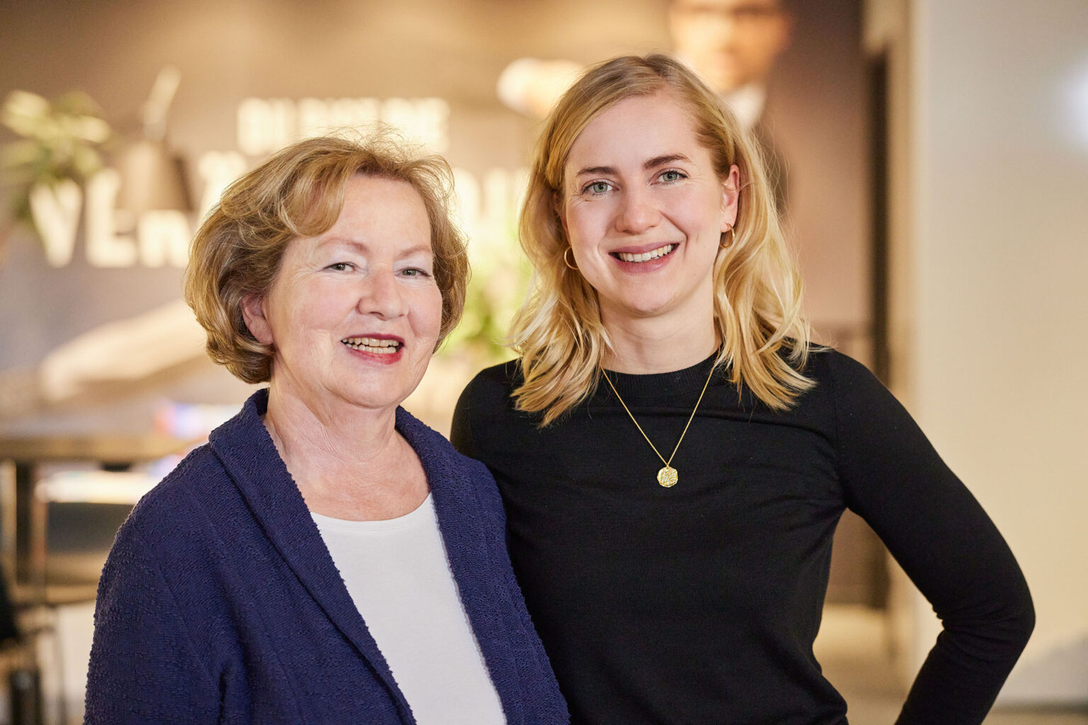 Eine ältere Frau mit kurzen, hellbraunen Haaren und eine jüngere Frau mit langen, blonden Haaren lächeln und stehen dicht beieinander in einem Innenraum. Die ältere Frau trägt eine blaue Strickjacke und ein weißes Oberteil, während die jüngere Frau ein schwarzes Oberteil und eine goldene Halskette trägt. Der Hintergrund ist unscharf und zeigt einige unklare Texte und eine Person in der Ferne.