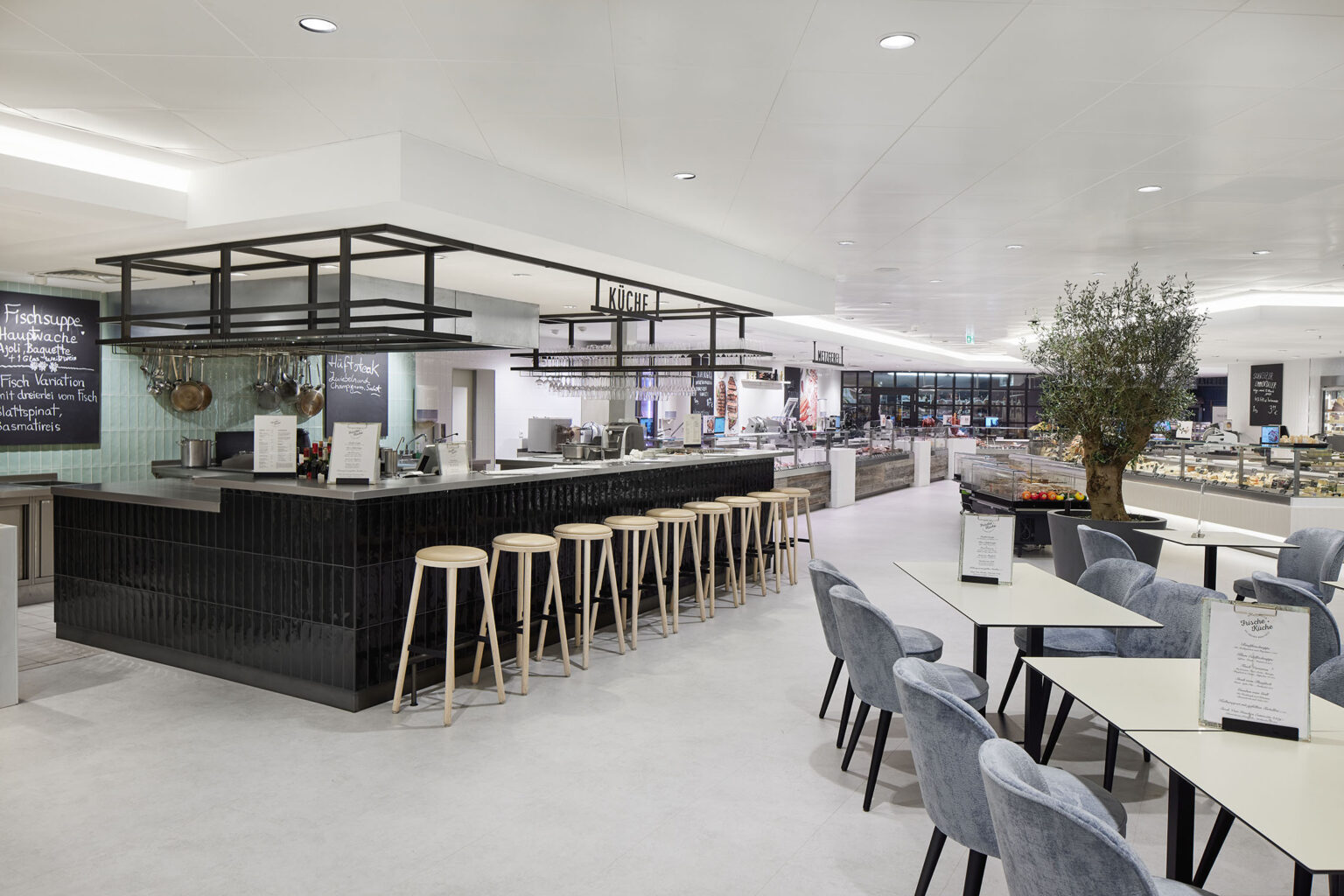 Ein moderner und heller Essbereich in einem Supermarkt, mit einer zentralen Küchentheke, die von Barhockern umgeben ist. Die Theke ist mit dunkelgrünen Kacheln verkleidet und bietet eine Auswahl an frischen Speisen. Links neben der Theke sind Menütafeln und Kochutensilien zu sehen. Im Vordergrund befinden sich Tische und Stühle, die zum Verweilen und Essen einladen. Im Hintergrund ist ein Olivenbaum als dekoratives Element platziert. Der Raum ist offen und geräumig gestaltet, mit viel natürlichem Licht und einem klaren, modernen Design.
