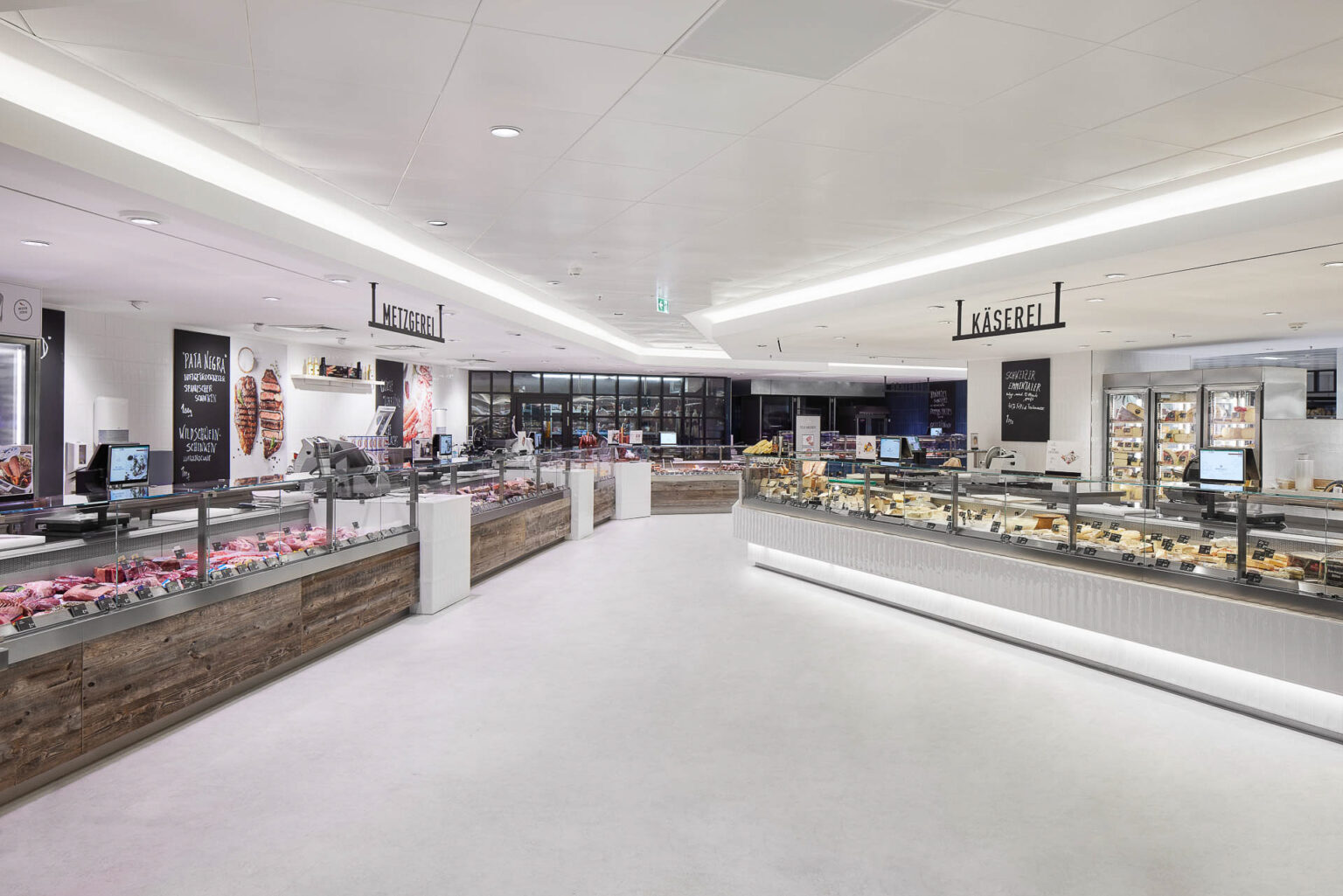 Ein großzügiger und heller Supermarktbereich mit verschiedenen Verkaufsstationen. Links ist eine Metzgerei-Theke zu sehen, die mit verschiedenen Fleischprodukten bestückt ist. Rechts befindet sich eine Käsetheke mit einer großen Auswahl an Käsesorten. Die Theken sind modern gestaltet, mit Glasabdeckungen und einer Mischung aus Holz- und Metallakzenten. Der Boden ist hell und der Raum gut beleuchtet. Im Hintergrund sind weitere Produkte und Regale zu erkennen, die eine Vielzahl von Lebensmitteln anbieten