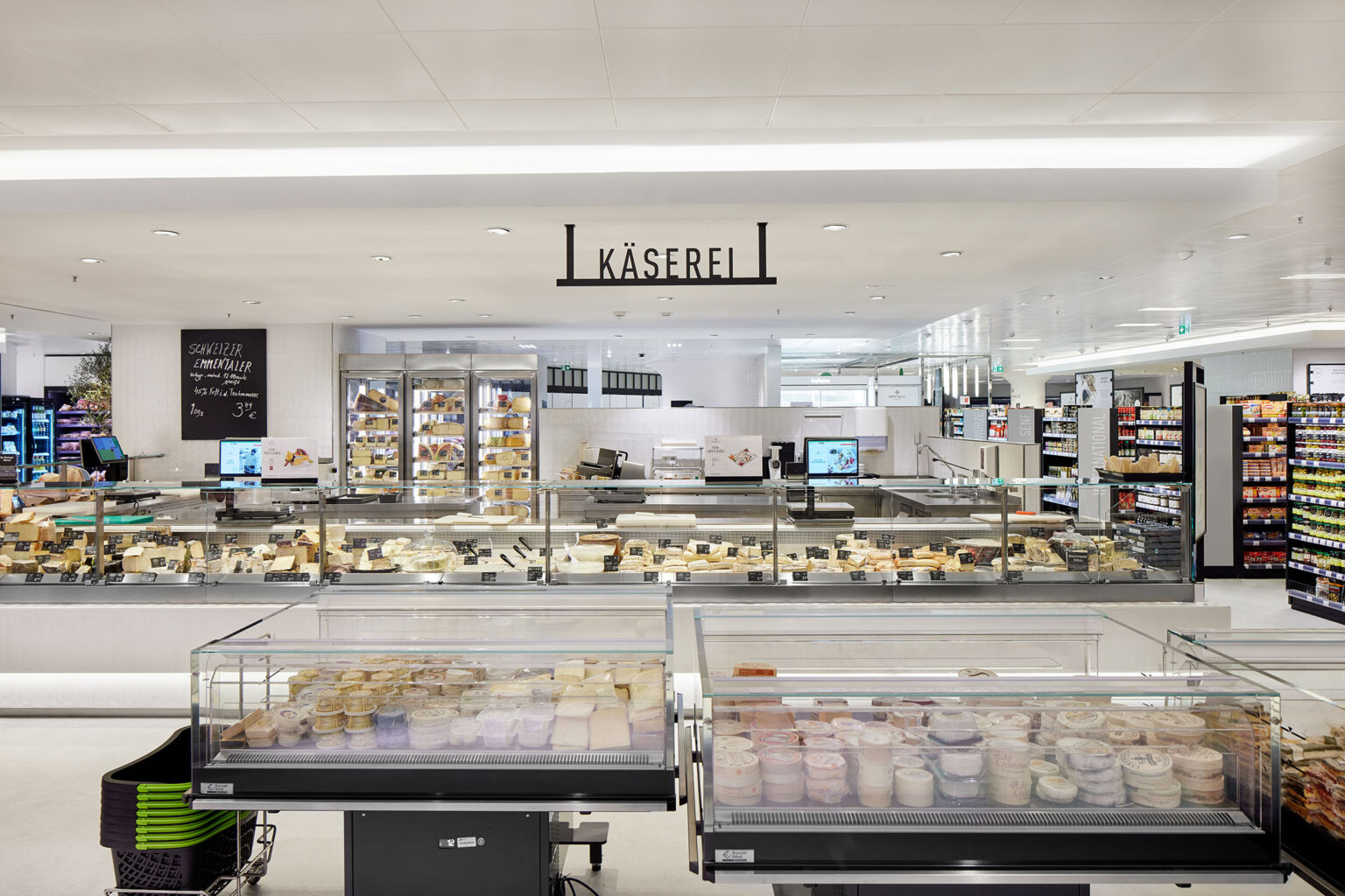 Ein großer und heller Supermarktbereich mit einer umfangreichen Käsetheke. Im Vordergrund sind Kühlvitrinen mit verschiedenen Käsesorten zu sehen. Dahinter befindet sich eine lange Theke mit einer noch größeren Auswahl an Käseprodukten. Über der Theke hängt ein Schild mit der Aufschrift 'Käserei'. Im Hintergrund sind weitere Regale mit zusätzlichen Produkten und Kühlvitrinen zu erkennen. Der Bereich ist sauber und modern gestaltet, mit weißem Boden und Decke sowie guter Beleuchtung, die die Produkte hervorhebt.