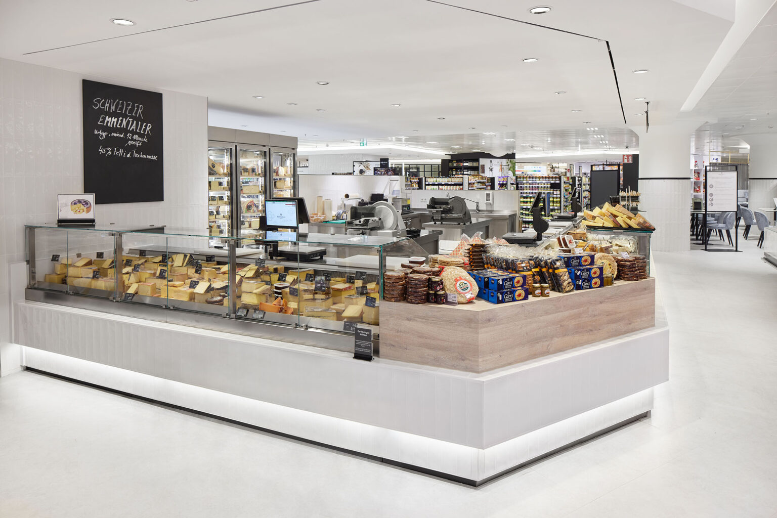 Ein moderner und heller Supermarktbereich mit einer Käsetheke im Vordergrund. Die Theke ist mit verschiedenen Käsesorten bestückt, die ordentlich hinter Glas präsentiert werden. Auf der rechten Seite der Theke befindet sich ein zusätzliches Display mit verschiedenen verpackten Käsesorten und Gebäck. An der Wand hinter der Theke hängt eine Tafel mit der Aufschrift 'Schweizer Emmentaler'. Der Raum ist sauber und gut beleuchtet, mit einem weißen Boden und einer modernen Einrichtung. Im Hintergrund sind weitere Bereiche des Supermarkts zu sehen.