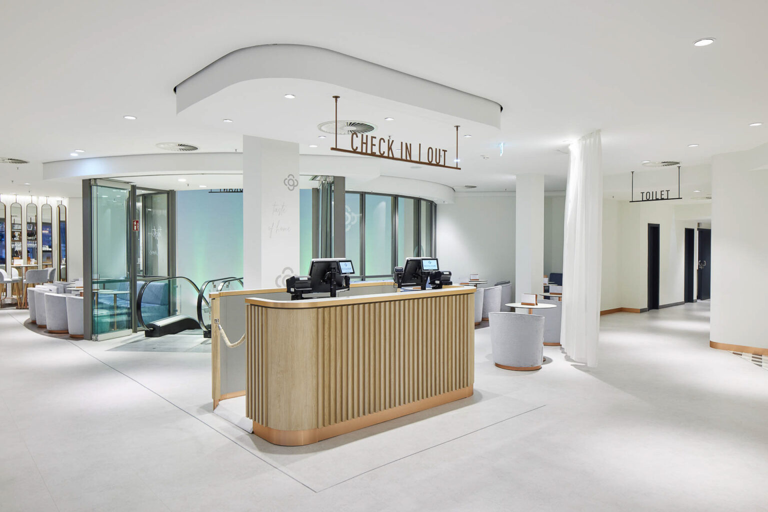 Ein moderner und heller Empfangsbereich in einem Gebäude. Eine Empfangstheke aus Holz steht im Vordergrund, ausgestattet mit zwei Check-in-Computern. Über der Theke hängt ein Schild mit der Aufschrift 'Check In | Out'. Im Hintergrund befinden sich ein Fahrstuhl und eine Treppe, die nach unten führen. Auf der rechten Seite ist ein weiteres Schild, das den Weg zu den Toiletten weist. Der Raum ist großzügig und minimalistisch gestaltet, mit hellen Wänden, einer weißen Decke und einem hellen Boden