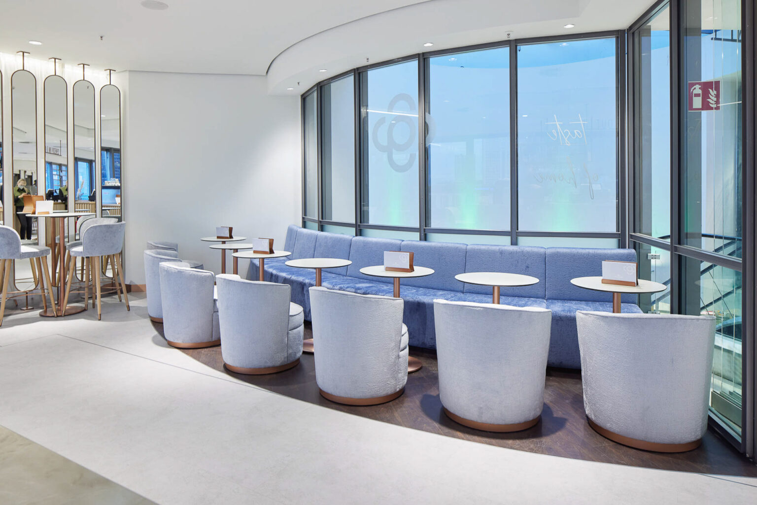 Ein stilvoller und heller Lounge-Bereich mit großen Fenstern, die viel natürliches Licht hereinlassen. Der Bereich ist mit einer langen blauen Sitzbank entlang der Fensterfront ausgestattet, vor der mehrere runde Tische und hellblaue Sessel stehen. Die Tische sind gleichmäßig verteilt, jeder mit einer kleinen Speisekarte oder einem Reservierungsschild darauf. Im Hintergrund sind hohe Spiegel und eine Theke mit Barhockern zu sehen. Die Einrichtung ist modern und elegant, mit einem Fokus auf Komfort und Ästhetik.