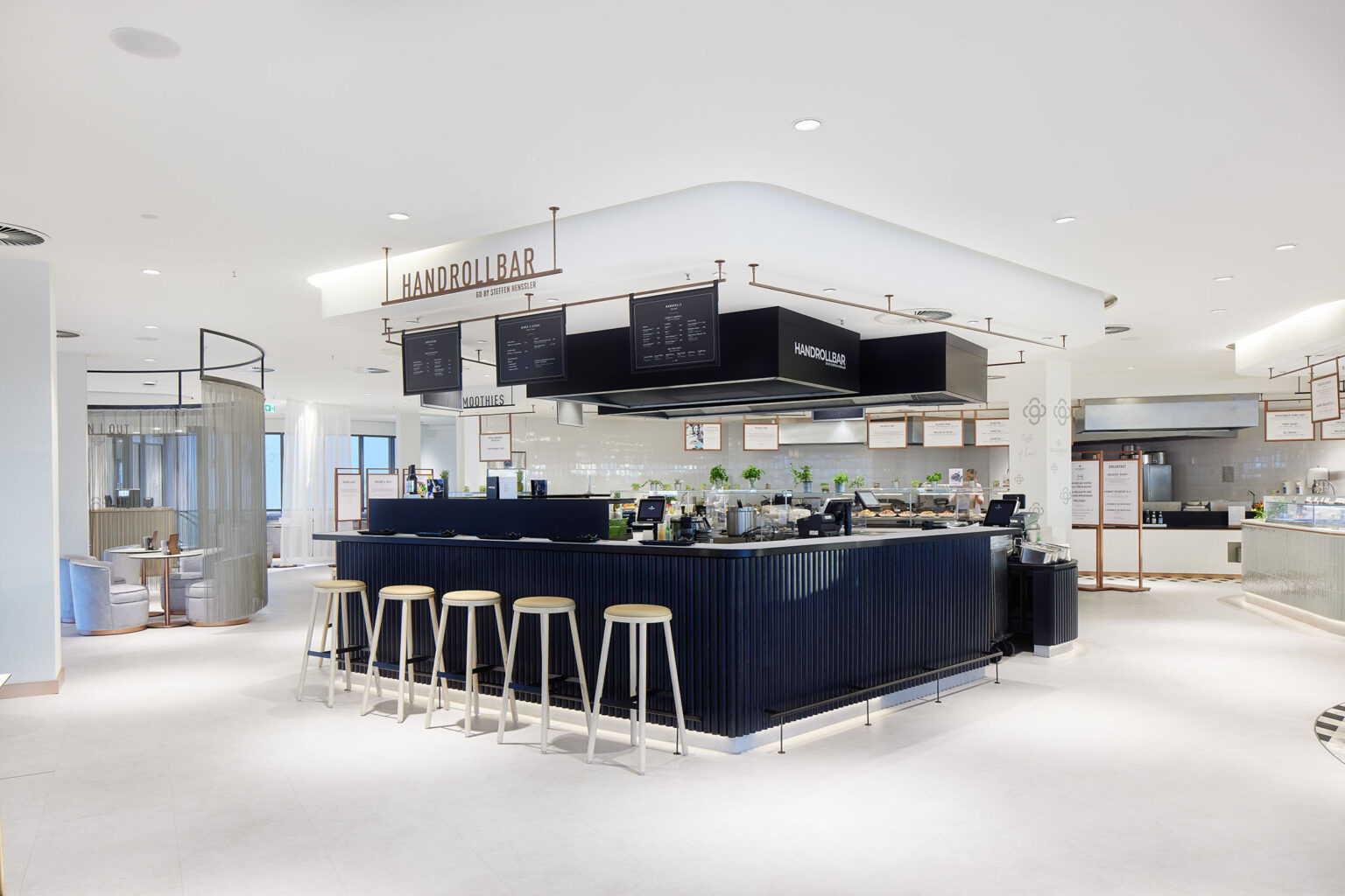 Ein moderner und heller Essbereich mit einer zentralen Theke, die als Handrollbar bezeichnet ist. Die Theke ist dunkelblau und von mehreren hellen Barhockern umgeben. Über der Theke hängen Schilder mit Menüs und der Aufschrift 'Handrollbar'. Im Hintergrund sind weitere Essensstationen und eine Theke mit verschiedenen Speisen und Getränken zu sehen. Der Raum ist offen und geräumig gestaltet, mit weißen Wänden und Böden sowie viel natürlichem Licht, das durch die großen Fenster auf der linken Seite hereinfällt.