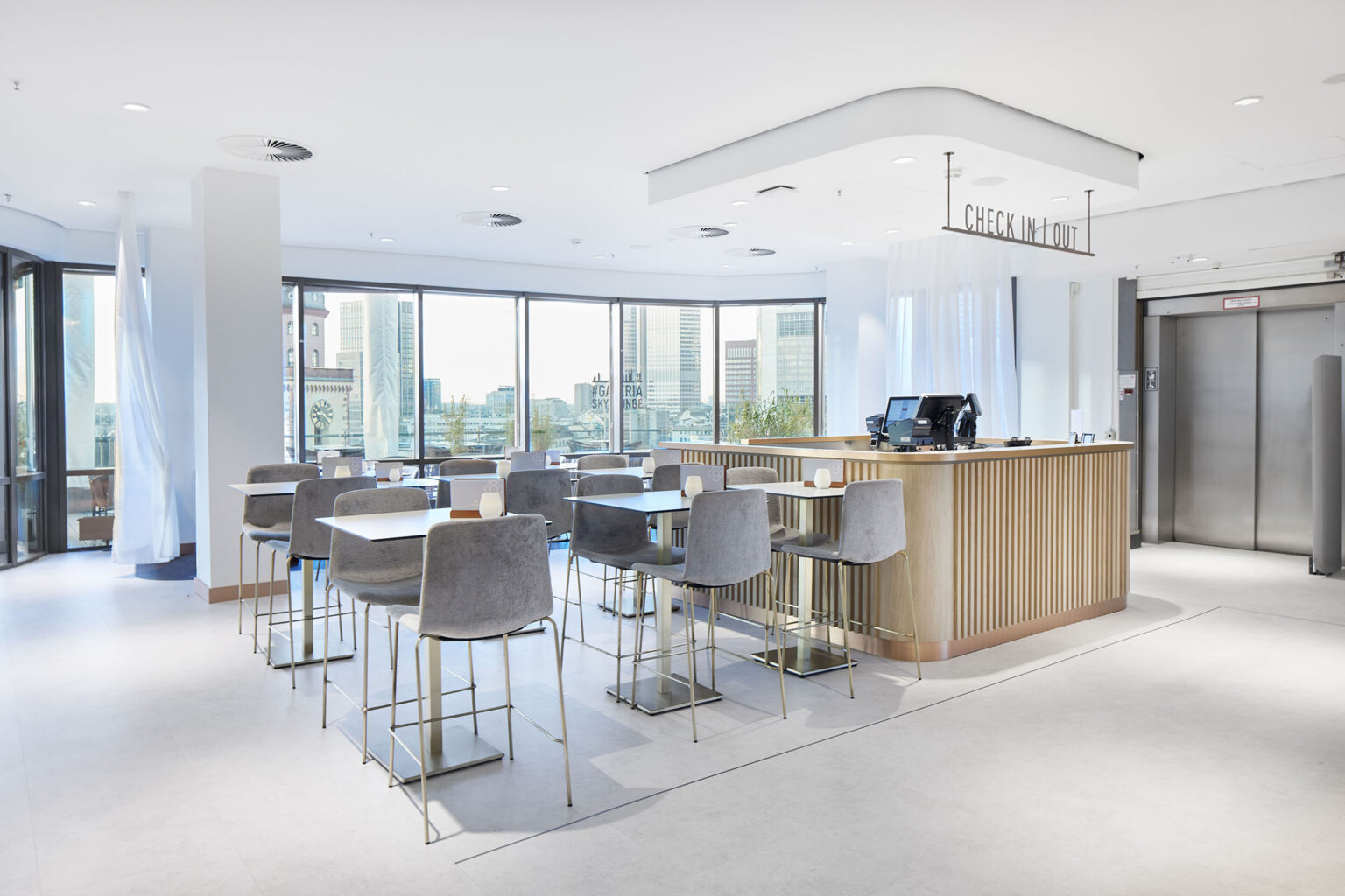 Ein moderner und heller Essbereich mit großen Fenstern, die eine Aussicht auf die Stadt bieten. In der Mitte des Raums befindet sich eine Empfangstheke aus Holz mit einem Schild darüber, das 'Check In | Out' anzeigt. Rund um die Theke stehen mehrere Tische und Stühle in einem eleganten grauen Design. Der Raum ist offen und einladend gestaltet, mit viel natürlichem Licht, das durch die Fenster strömt. Auf der rechten Seite befinden sich Aufzüge, die den Zugang zu anderen Etagen ermöglichen.