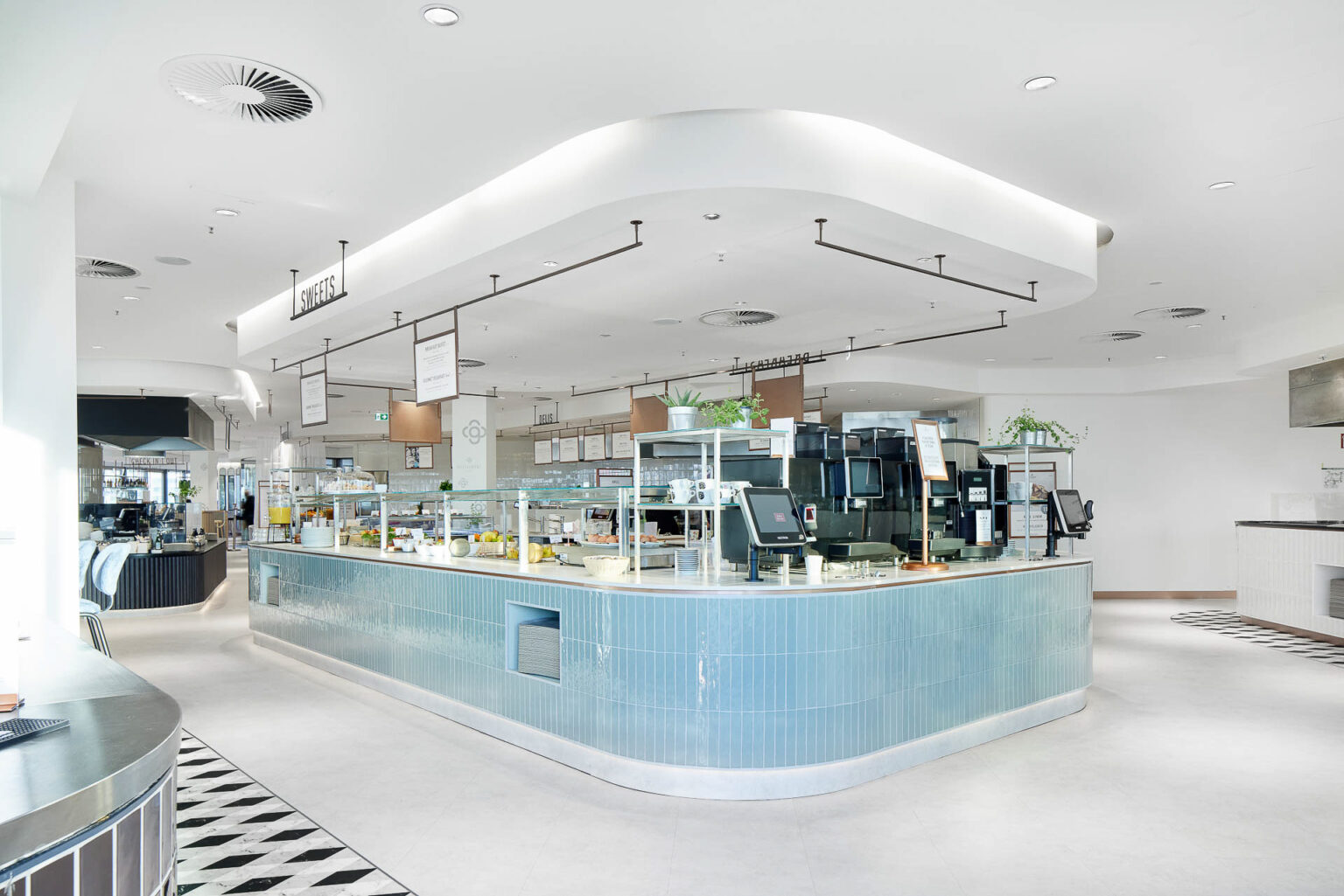 Ein modernes und helles Selbstbedienungsrestaurant mit einer zentralen Theke in einem geschwungenen Design. Die Theke ist mit hellblauen Kacheln verkleidet und mit verschiedenen Speisen und Getränken bestückt. Über der Theke hängen Schilder, die die verschiedenen Essensbereiche wie 'Sweets' kennzeichnen. Der Raum ist großzügig gestaltet, mit viel natürlichem Licht und Pflanzen zur Dekoration. Die Einrichtung ist modern und sauber, mit einem weißen Boden und Decke sowie stilvollen schwarzen und weißen Fliesen.