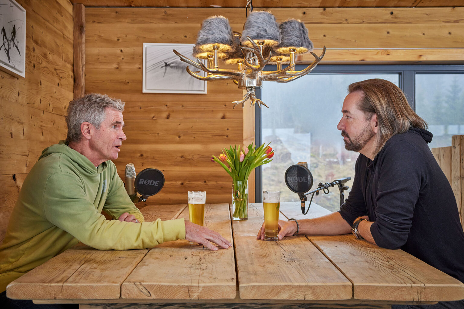 Zwei Männer sitzen an einem Holztisch in einem gemütlichen, holzgetäfelten Raum. Jeder von ihnen hat ein Glas Bier vor sich. Der Mann links trägt einen grünen Hoodie, während der Mann rechts einen schwarzen Pullover trägt. Auf dem Tisch stehen zwei Mikrofone mit der Aufschrift 'RØDE', was darauf hindeutet, dass sie eine Unterhaltung oder ein Interview führen. In der Mitte des Tisches steht eine Vase mit bunten Tulpen. Über ihnen hängt ein dekorativer Kronleuchter mit Hirschgeweihen und pelzbesetzten Lampenschirmen. Durch das Fenster im Hintergrund ist eine neblige Landschaft zu sehen.