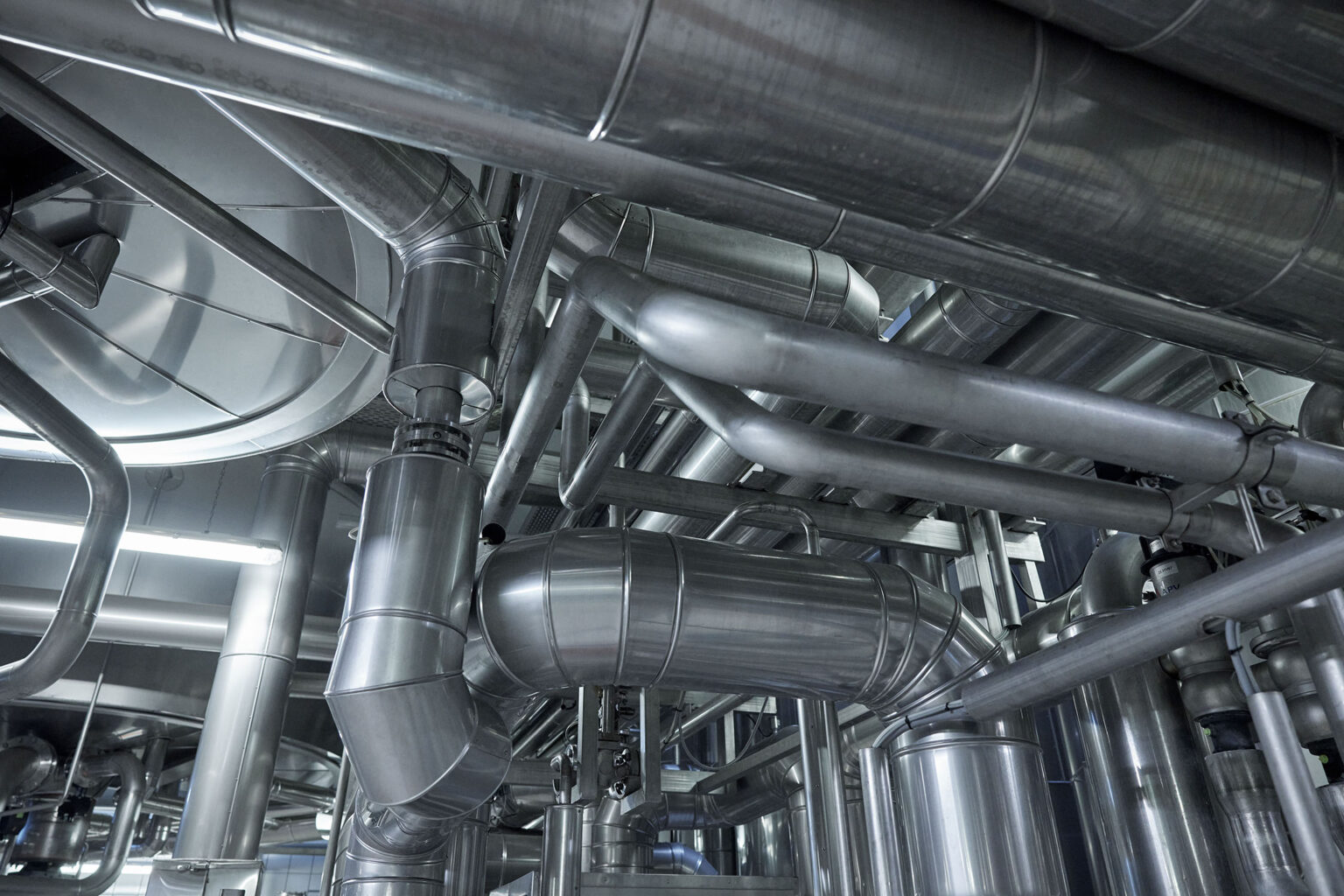 Ein dichtes Netzwerk aus Edelstahlrohren und -leitungen in einer industriellen Anlage. Die Rohre verlaufen in verschiedenen Richtungen und sind stabil an der Decke und an den Wänden montiert. Die glänzenden Metallrohre reflektieren das Licht, was die Sauberkeit und die moderne Beschaffenheit der Anlage betont. Die gesamte Struktur wirkt robust und gut organisiert, was auf eine effiziente industrielle Umgebung hinweist.
