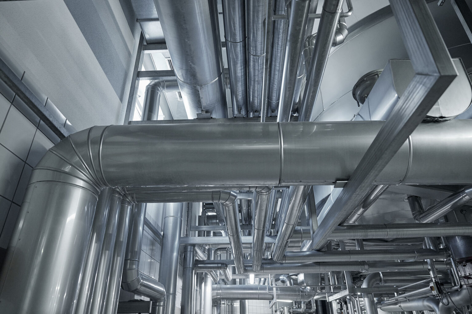 Detailansicht eines Netzwerks von Edelstahlrohren und -leitungen in einer industriellen Anlage. Die Rohre verlaufen in verschiedenen Richtungen und sind an der Decke und den Wänden befestigt. Die glänzende Oberfläche der Rohre reflektiert das Licht, was die moderne und saubere Umgebung der Anlage betont. Diese Struktur vermittelt den Eindruck von Effizienz und Organisation in der industriellen Umgebung.