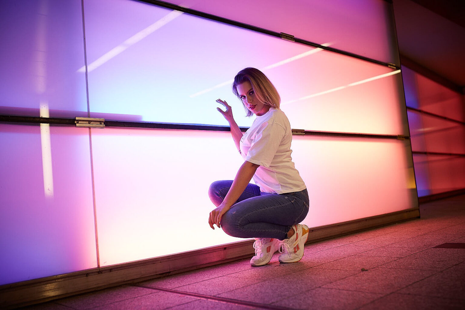 Eine junge Frau in einem lässigen Outfit posiert vor einer beleuchteten Wand, die in wechselnden Pink- und Lilatönen leuchtet. Sie hockt sich hin, eine Hand an der Wand, und dreht ihren Kopf zur Kamera, wobei ein halbes Lächeln auf ihrem Gesicht zu sehen ist. Ihr weißes T-Shirt kontrastiert mit dem farbenfrohen Hintergrund, und ihre Blue Jeans und weißen Sneaker komplettieren den entspannten Look. Die Szene strahlt eine urbane, trendige Atmosphäre aus.