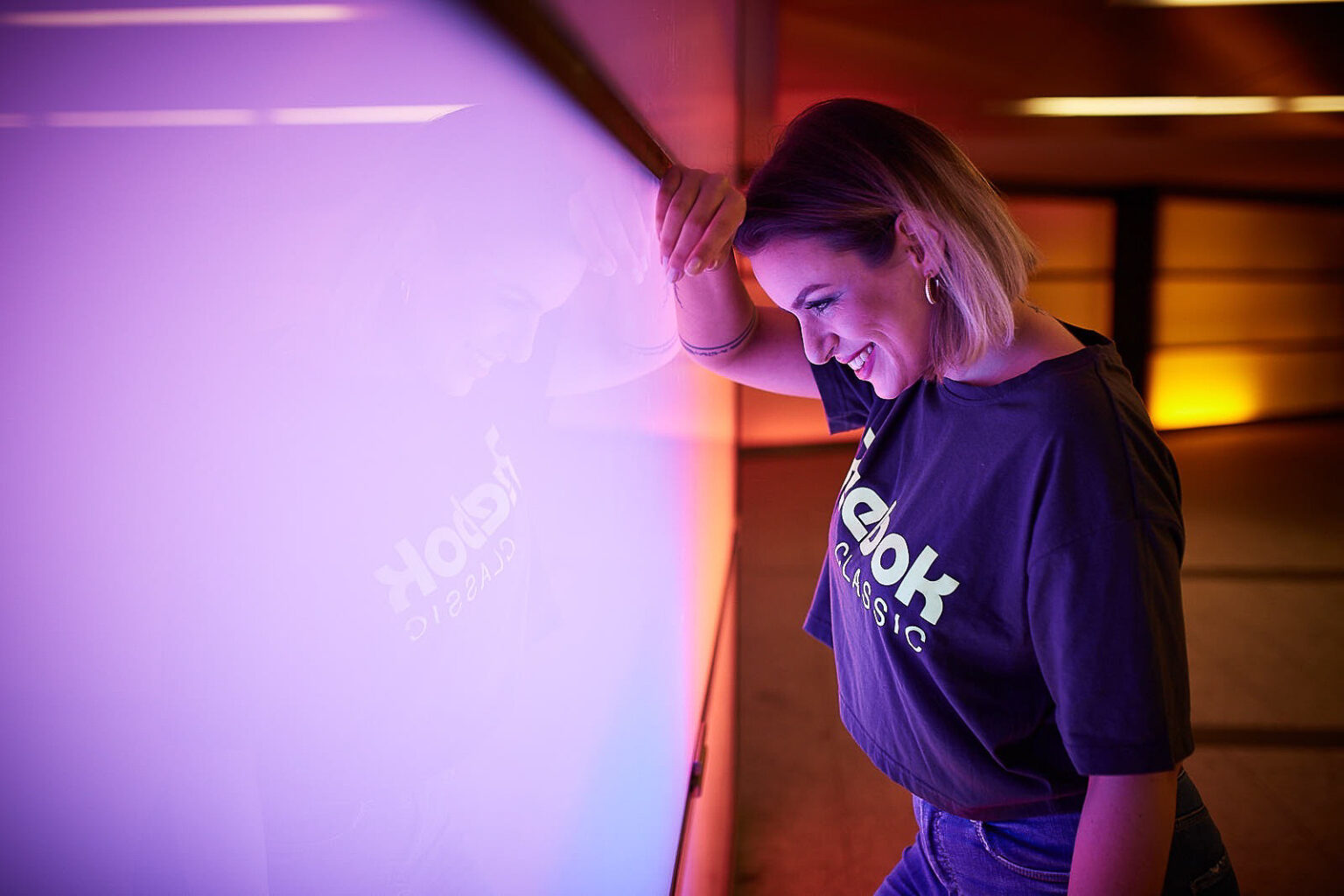 Eine junge Frau mit einem strahlenden Lächeln lehnt sich in einem Raum mit lebhafter Beleuchtung gegen eine lila beleuchtete Wand. Sie trägt ein dunkles T-Shirt mit dem Logo "Reebok Classic" und schaut nach unten, während die Umgebungslichter ihre Umrisse hervorheben und eine warme und freudige Atmosphäre schaffen. Ihr Spiegelbild ist sanft auf der glänzenden Oberfläche hinter ihr zu erkennen, was der Szene eine leichte und spielerische Stimmung verleiht.