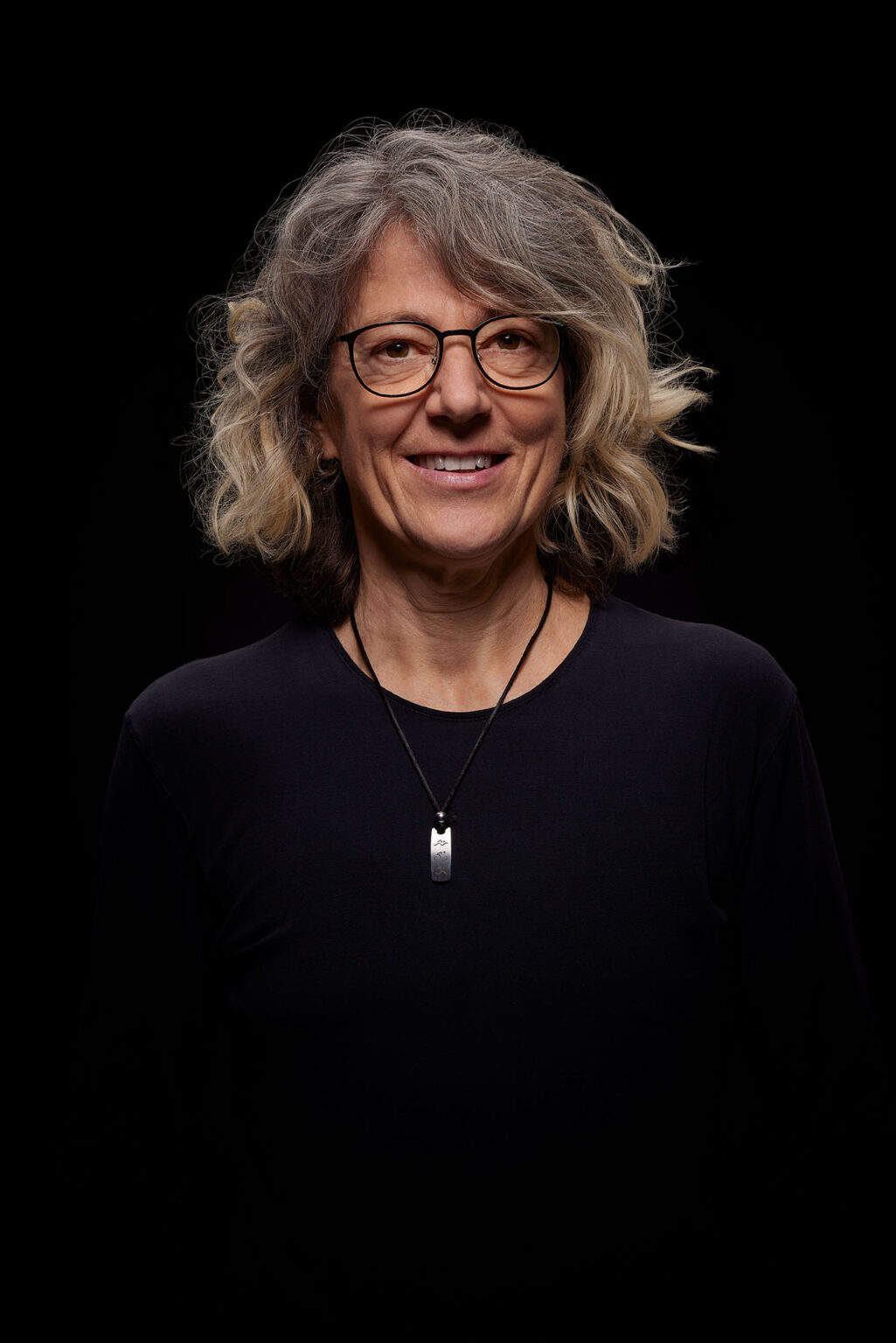 Ein Porträt einer älteren Frau vor einem schwarzen Hintergrund. Sie trägt eine Brille und ein schwarzes Oberteil. Um ihren Hals hängt eine Kette mit einem rechteckigen Anhänger. Die Frau hat graue, lockige Haare und lächelt leicht in die Kamera.