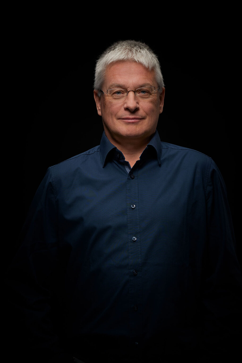 Ein Porträt eines älteren Mannes vor einem schwarzen Hintergrund. Er trägt eine Brille und ein dunkelblaues Hemd. Der Mann hat kurze, graue Haare und schaut direkt in die Kamera mit einem leichten Lächeln.