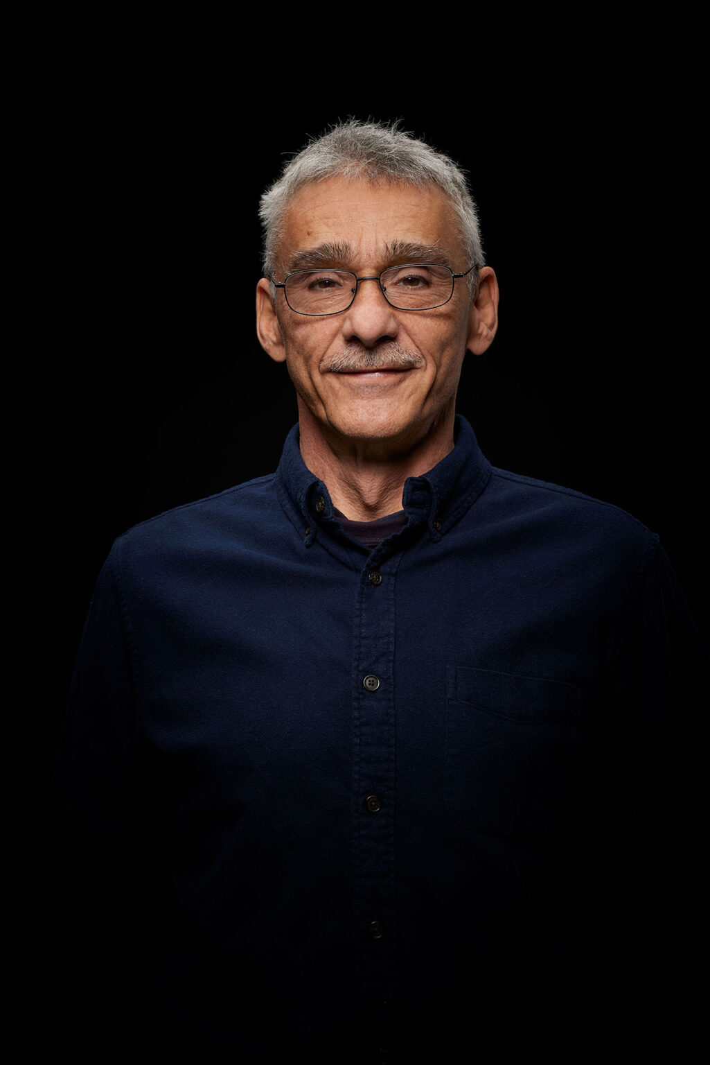 Ein Porträt eines älteren Mannes vor einem schwarzen Hintergrund. Er trägt eine Brille und ein dunkelblaues Hemd. Der Mann hat graue, kurze Haare und einen Schnurrbart. Er schaut direkt in die Kamera und lächelt leicht.