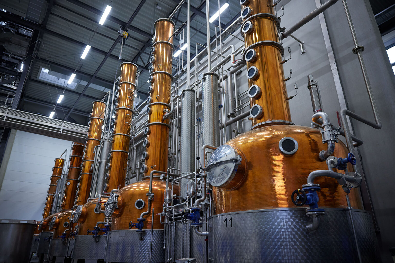 Eine industrielle Anlage mit mehreren großen, kupferfarbenen Destillationstürmen in einer Reihe. Die Türme sind mit zahlreichen Rohren, Ventilen und Leitungen verbunden. Im Vordergrund sind die unteren Teile der Destillationstürme zu sehen, die mit Edelstahlbehältern ausgestattet sind. Die Anlage befindet sich in einem großen, gut beleuchteten Raum mit einem hohen Metall-Dach.