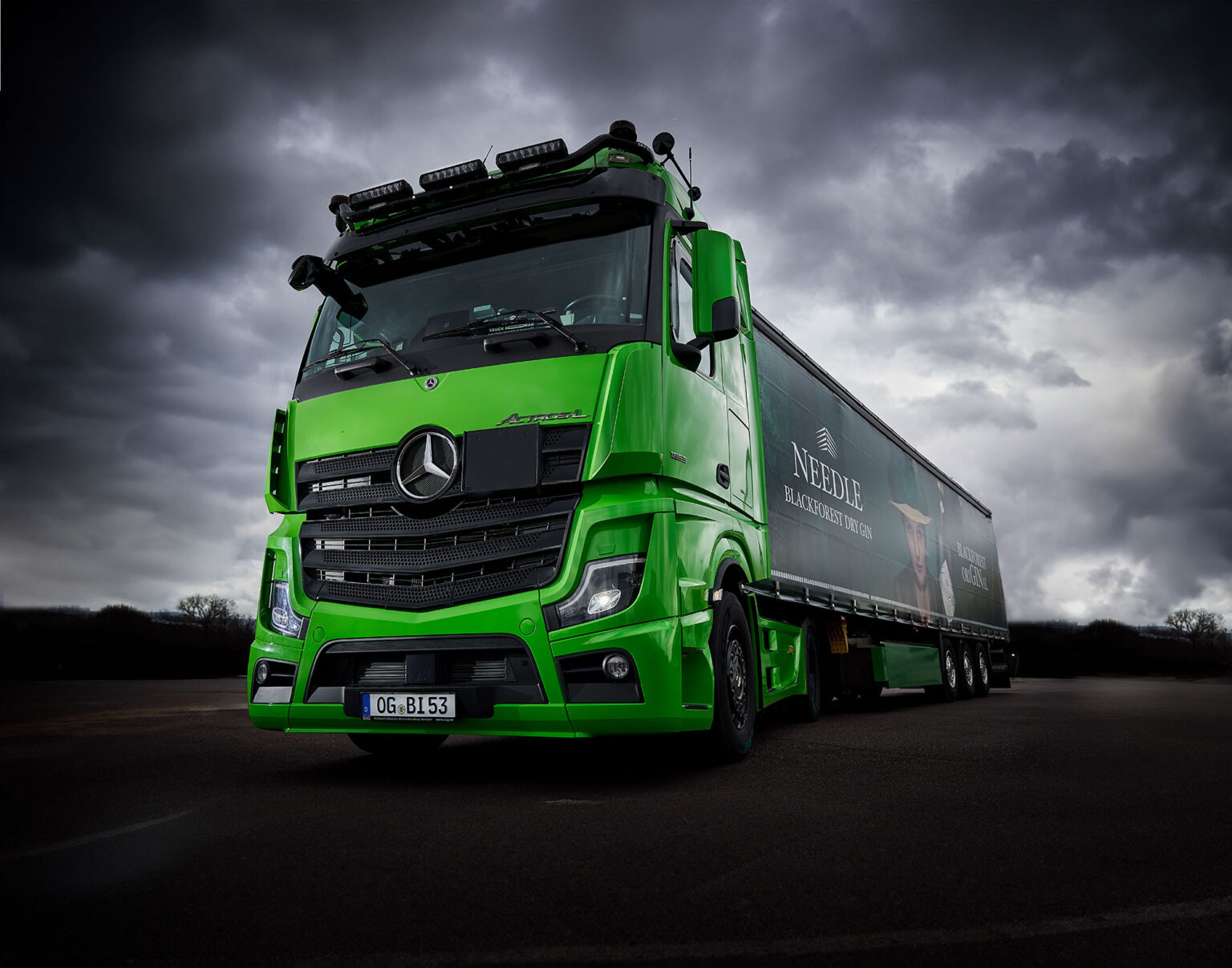 Ein großer grüner Lastwagen der Marke Mercedes-Benz steht auf einer Straße unter einem dramatisch bewölkten Himmel. Der Lastwagen hat die Aufschrift 'NEEDLE BLACK FOREST DRY GIN' auf dem Anhänger und trägt ein deutsches Nummernschild. Die Szenerie ist düster und betont die kräftigen Farben des Fahrzeugs vor dem dunklen Hintergrund
