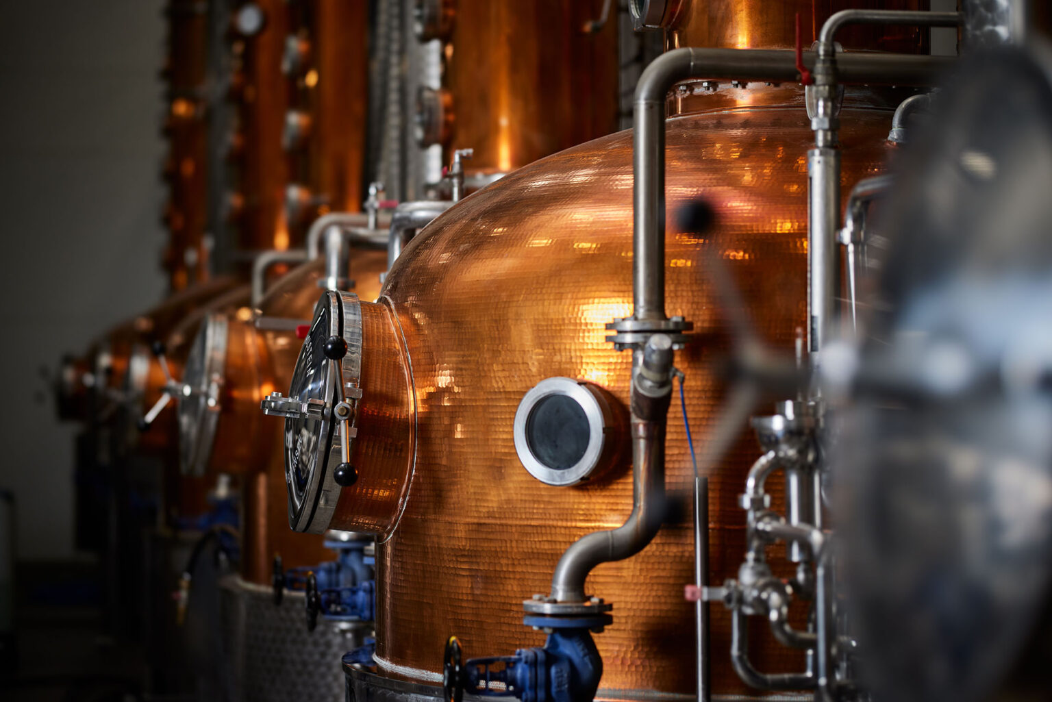 Ein Innenraum mit mehreren großen, kupfernen Destillationsapparaten in einer Brennerei. Die Apparate sind mit verschiedenen Rohren und Ventilen ausgestattet und reflektieren das Licht. Die Szene wirkt technisch und industriell.