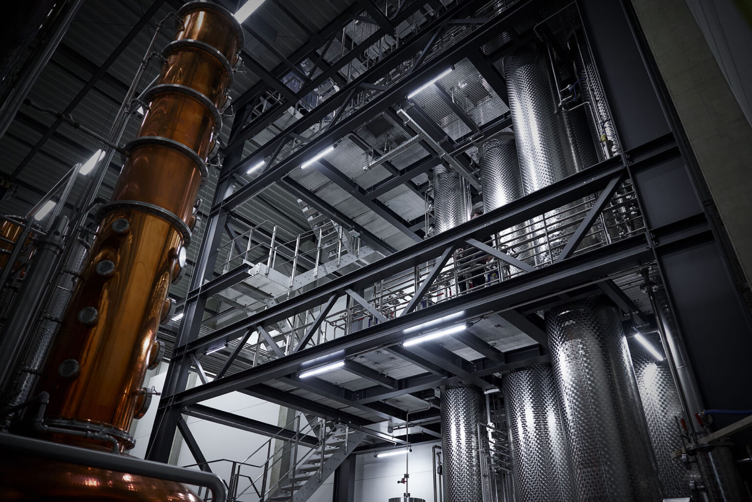 Eine industrielle Anlage mit mehreren Etagen aus Metallstrukturen und Plattformen. Links im Bild ist ein großer, kupferfarbener Destillationsturm zu sehen. Rechts befinden sich mehrere große, zylindrische Edelstahltanks. Die Anlage ist gut beleuchtet und zeigt zahlreiche Rohre und Treppen, die für den Zugang zu verschiedenen Ebenen und Maschinen verwendet werden