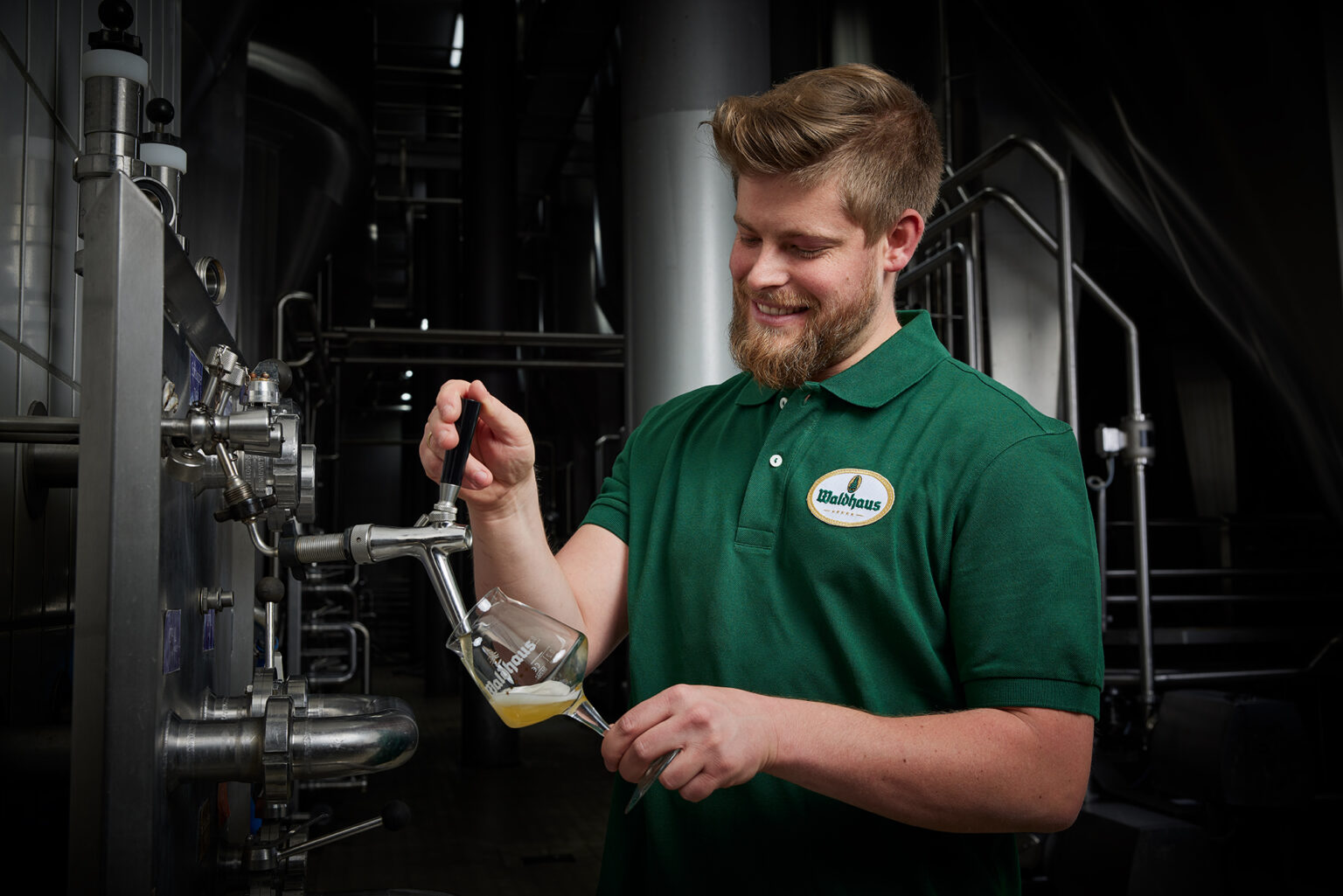 Ein junger Mann mit Bart und kurzen Haaren in einem grünen Poloshirt mit dem Logo von 'Waldhaus' zapft ein Glas Bier in einer Brauerei. Er lächelt und schaut auf das Glas, während er den Zapfhahn bedient. Im Hintergrund sind industrielle Brauereiausrüstungen zu sehen.