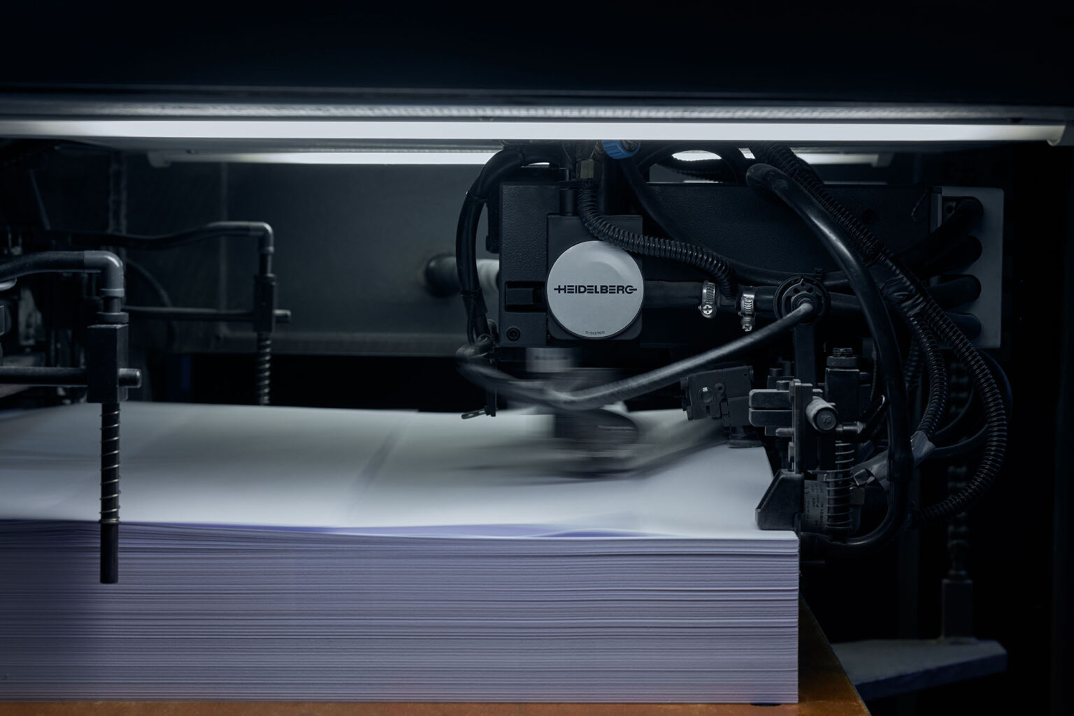 Eine Nahaufnahme einer industriellen Druckmaschine der Marke Heidelberg in Betrieb. Die Maschine verarbeitet einen Stapel weißer Papierbögen, die durch mechanische Arme und Greifer schnell bewegt werden. Die Bewegungsunschärfe zeigt die hohe Geschwindigkeit, mit der die Papierbögen durch die Maschine geführt werden. Die Beleuchtung und die zahlreichen Kabel und Schläuche der Maschine sind ebenfalls sichtbar.