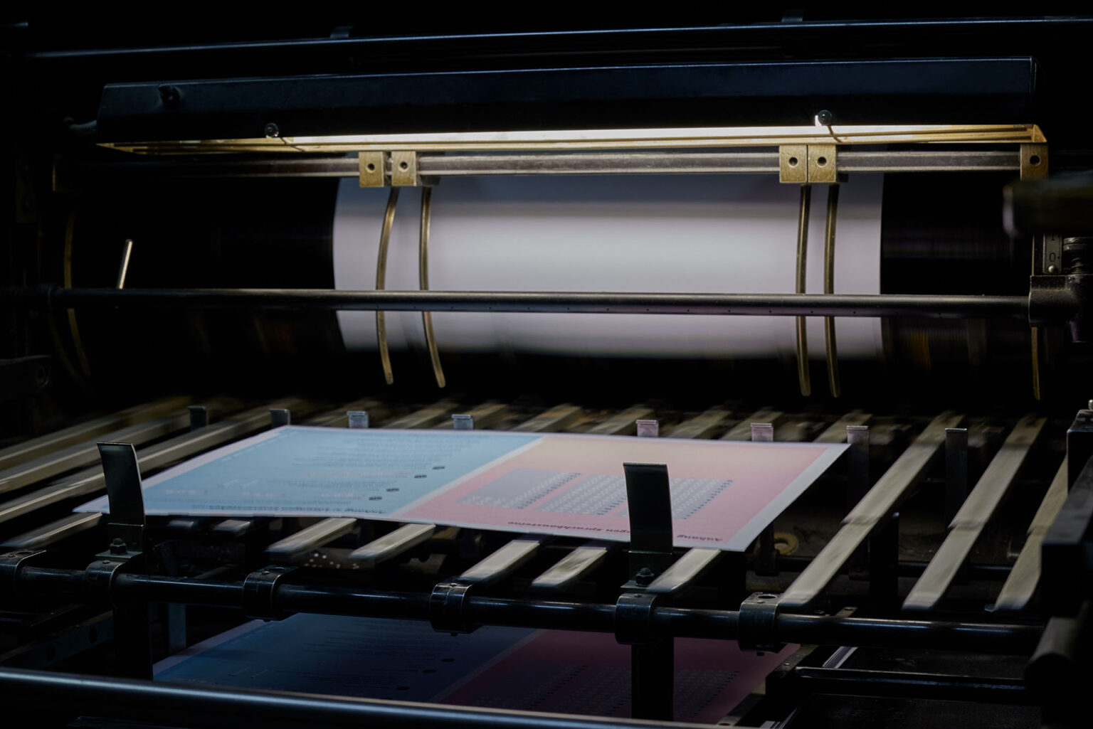 Eine Nahaufnahme einer industriellen Druckmaschine in Betrieb, die farbige Drucke produziert. Ein weißer Papierbogen wird durch die Maschine geführt und durchläuft mehrere Walzen und Metallstangen. Ein bereits bedruckter Bogen mit Text und Grafiken ist im Vordergrund zu sehen. Die Druckmaschine ist mit Beleuchtung ausgestattet, die den Druckprozess gut sichtbar macht.