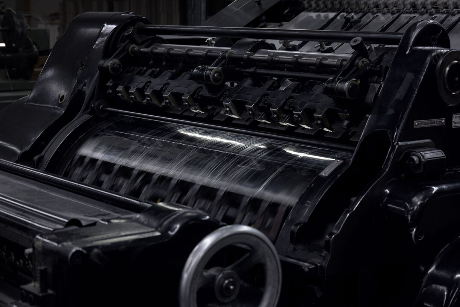 Eine Nahaufnahme einer großen, industriellen Druckmaschine in Betrieb. Die Maschine hat zahlreiche rotierende Walzen und Zahnräder, die Papier durch den Druckprozess befördern. Die schwarzen Metallteile der Maschine sind robust und zeigen Anzeichen von intensivem Gebrauch. Die Bewegung der Walzen ist in verschwommenen Linien sichtbar, was die Geschwindigkeit und den kontinuierlichen Betrieb der Maschine zeigt.